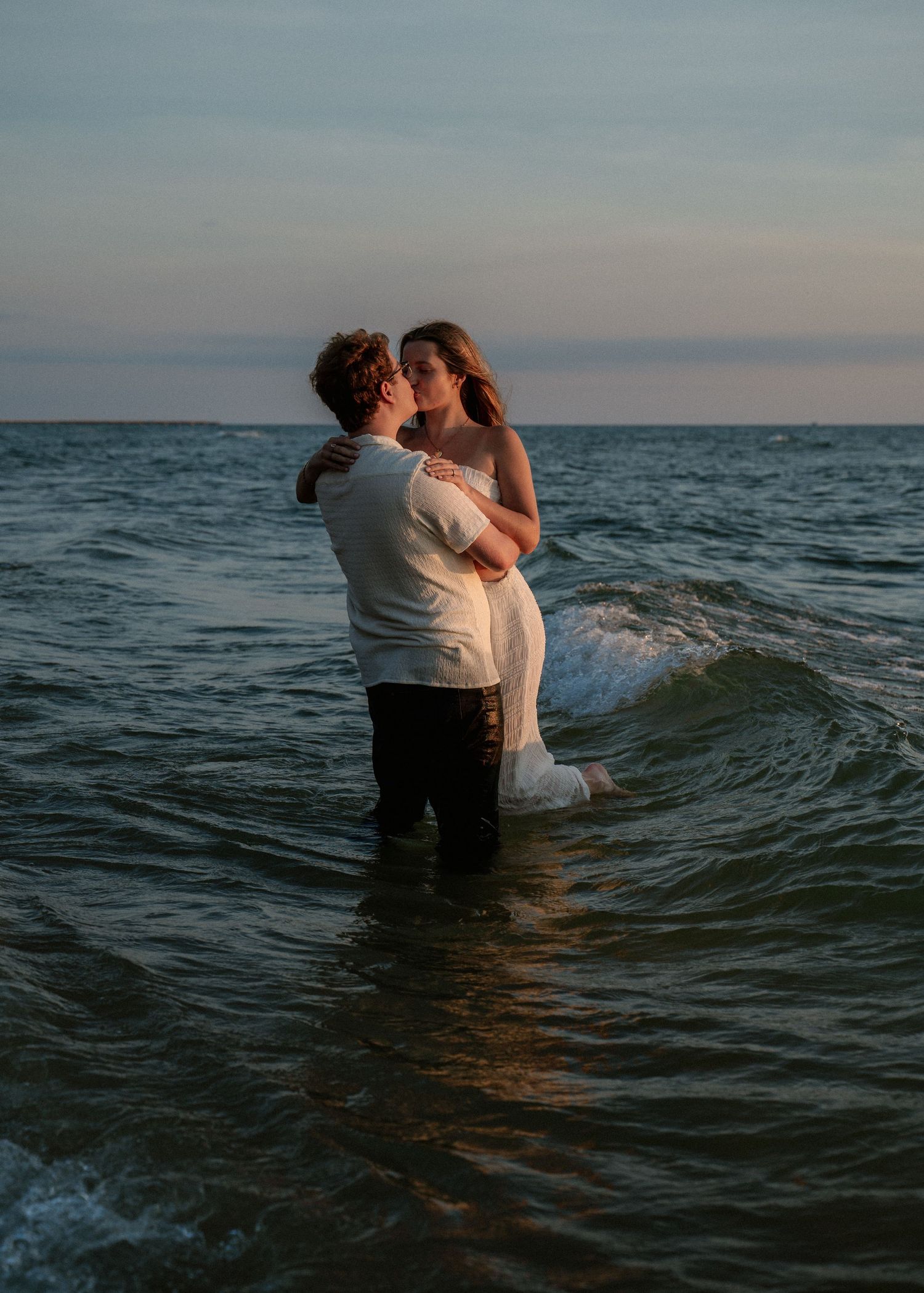 Couple sharing romantic moments in ocean waves during sunset photoshoot.