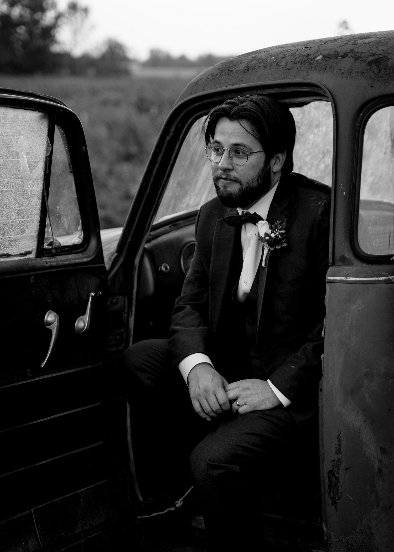 A groom in a black tuxedo sits in a vintage car for a moody black and white portrait.