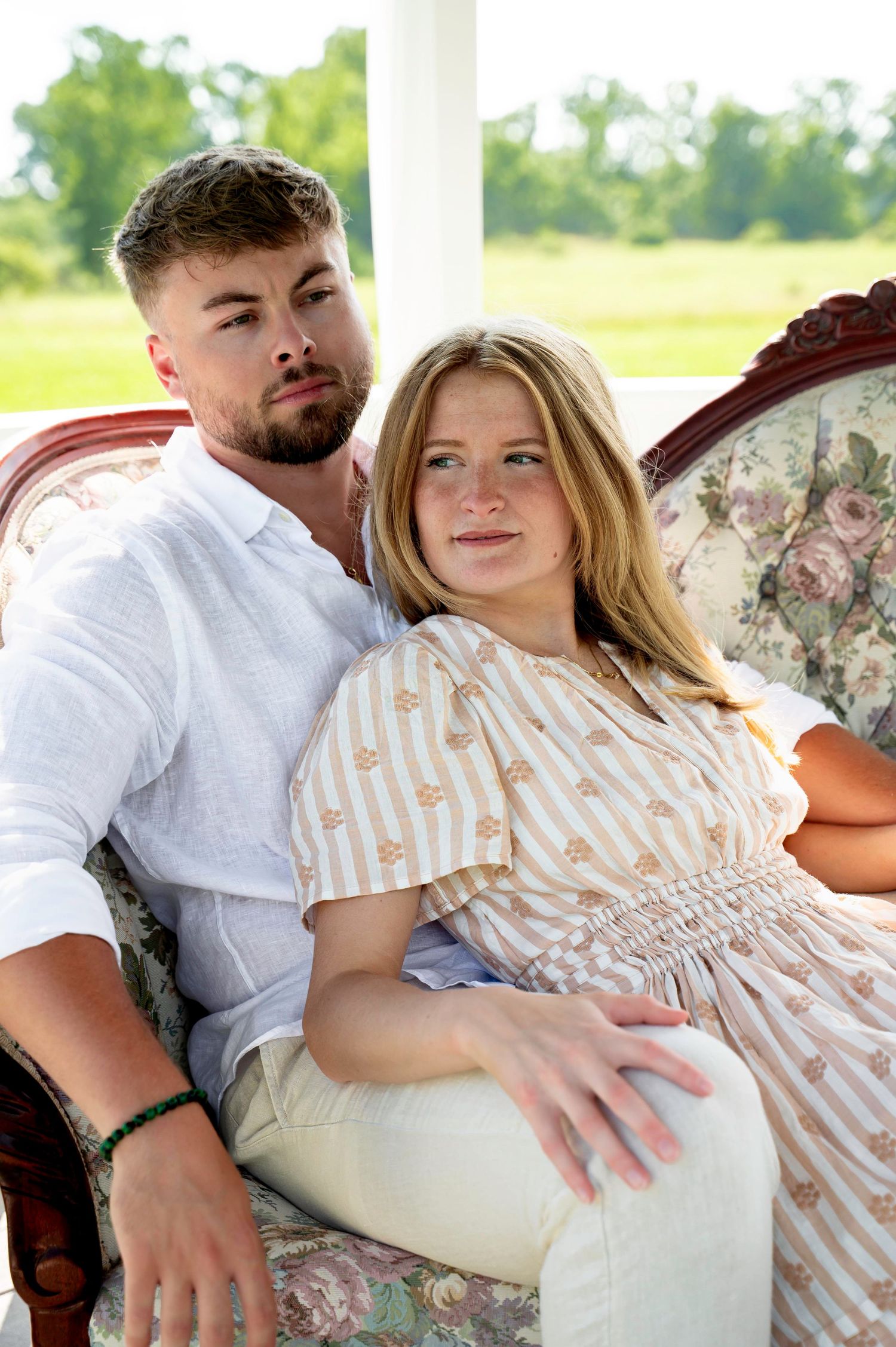 Couple lounging on antique floral sofa in bright sunlit room.