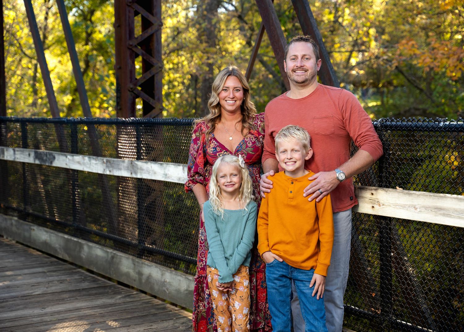 Family poses playfully on wooden bridge with fall colors and water in background.