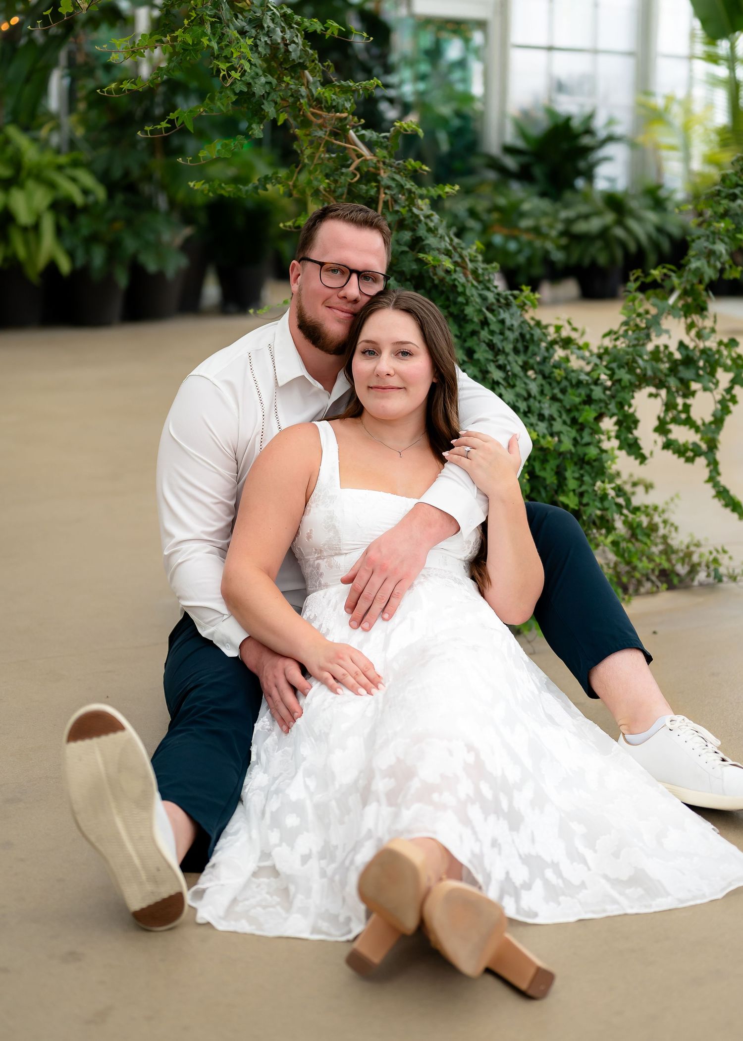 A couple relaxes together in casual attire on a light colored floor with greenery visible in the background.