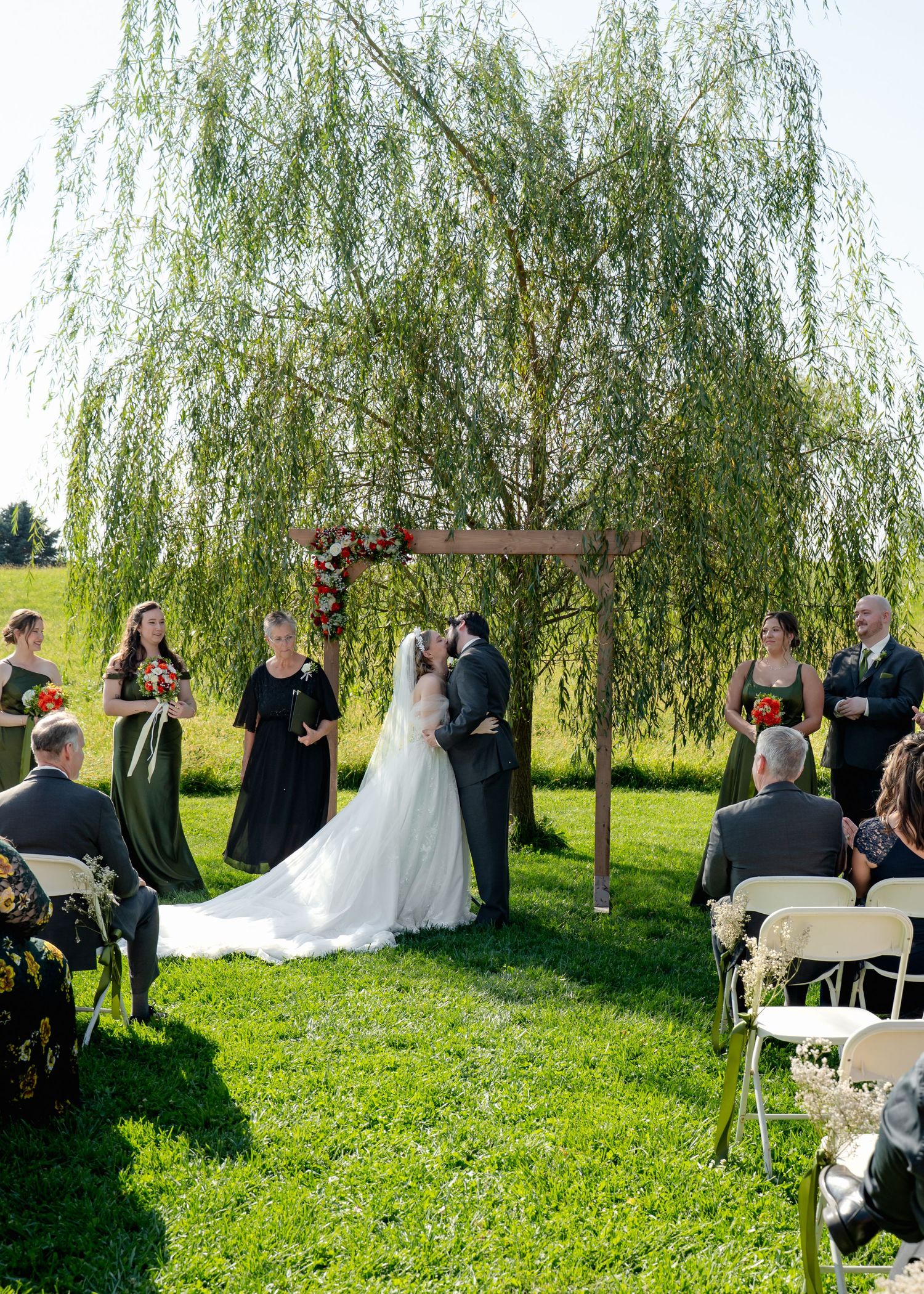 Wedding ceremony in progress under a willow tree with guests seated on folding chairs.