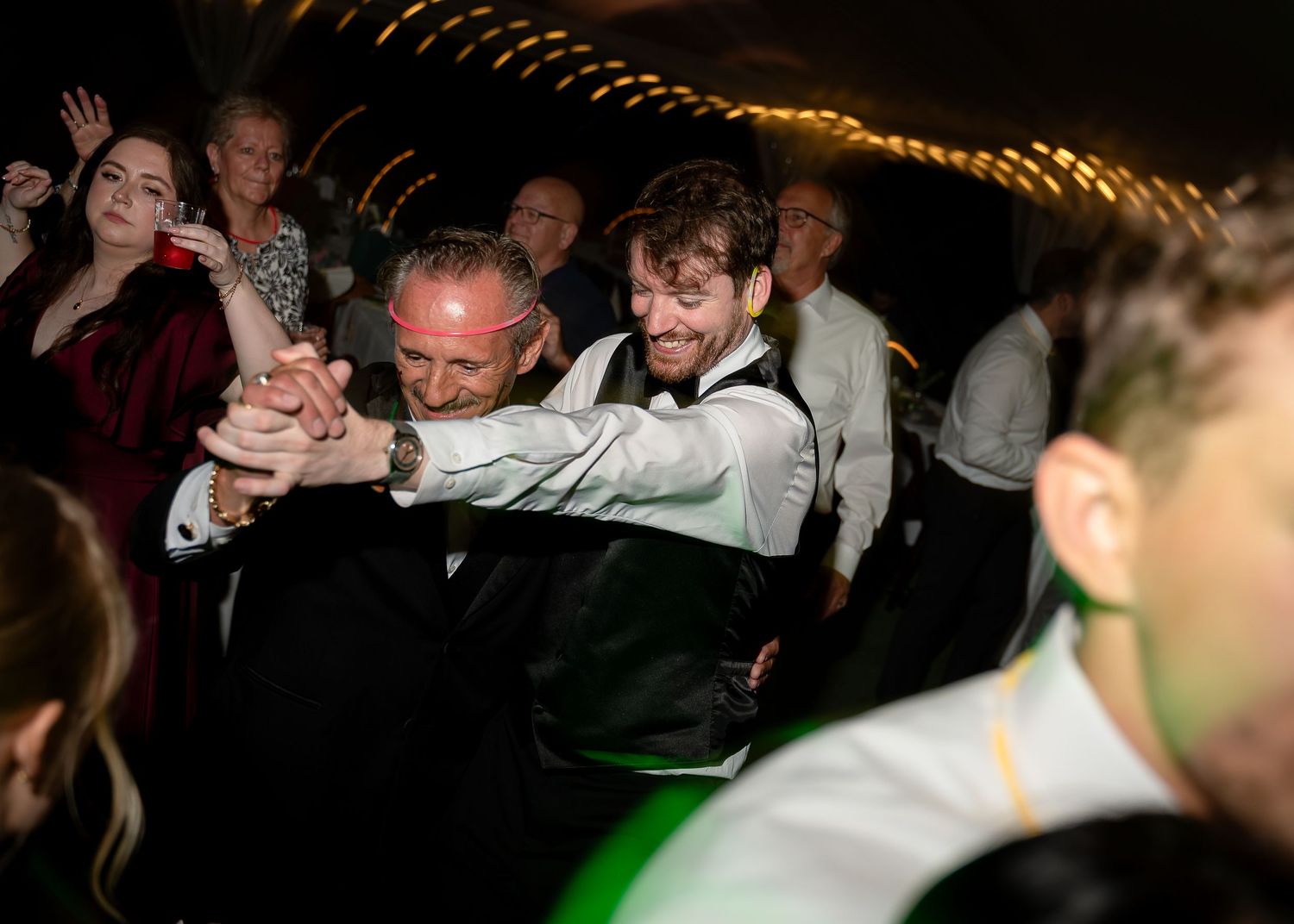 Two men dancing energetically at a lively wedding reception party with guests celebrating in the background.