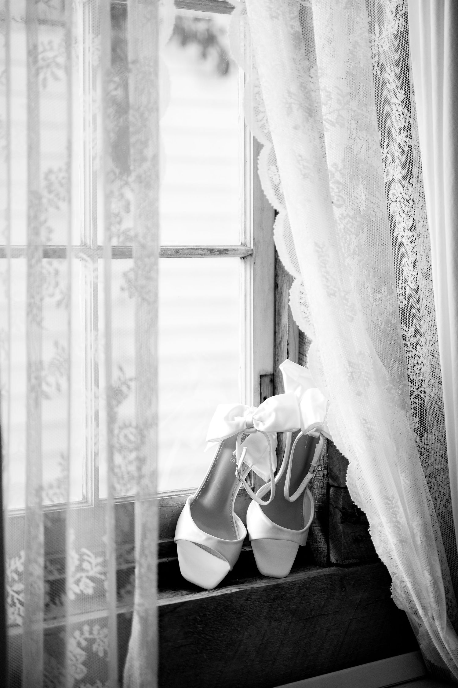 Black and white photo of white bridal heels with ribbon ties resting on a windowsill with lace curtains.