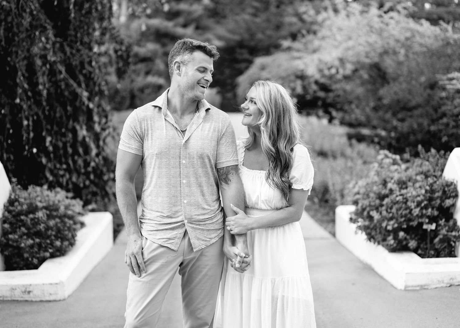 Black and white photo of a smiling couple walking together in a lush garden, woman in white dress.
