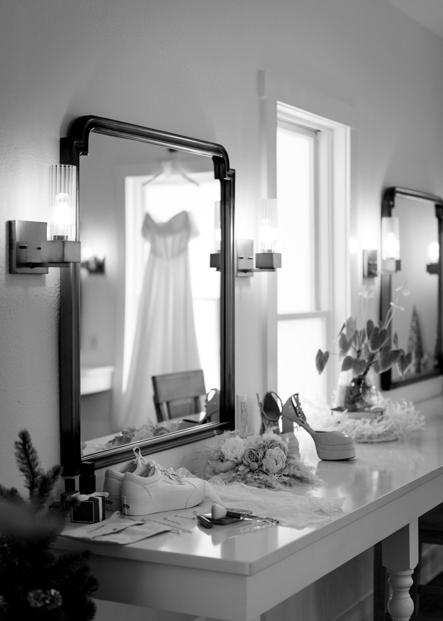 Black and white photo of a bridal vanity with wedding dress reflected in mirror, bouquet, and heels on display.