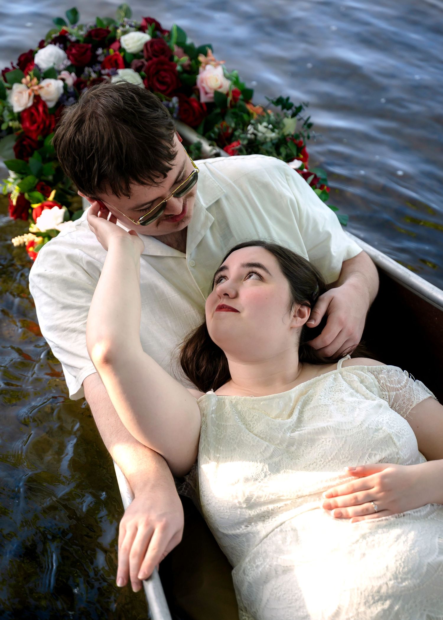 Couple in a boat surrounded by flowers, woman lying back gazing up at man in a romantic outdoor scene.