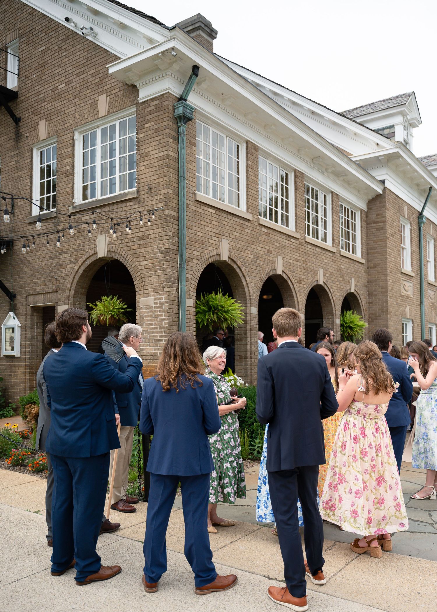 Guests in formal attire gather outside an elegant brick building with arched entrances for an outdoor event.