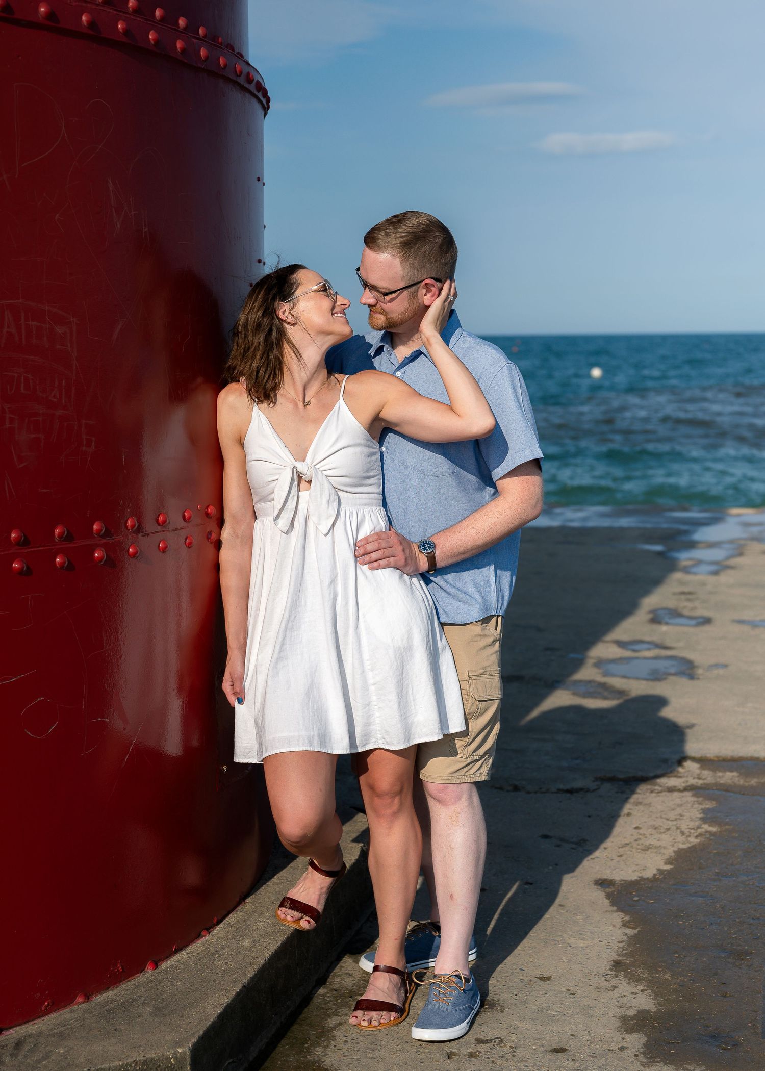 Couple smiling at each other near a red lighthouse on a sunny day by the ocean, woman in white dress, man in blue shirt.