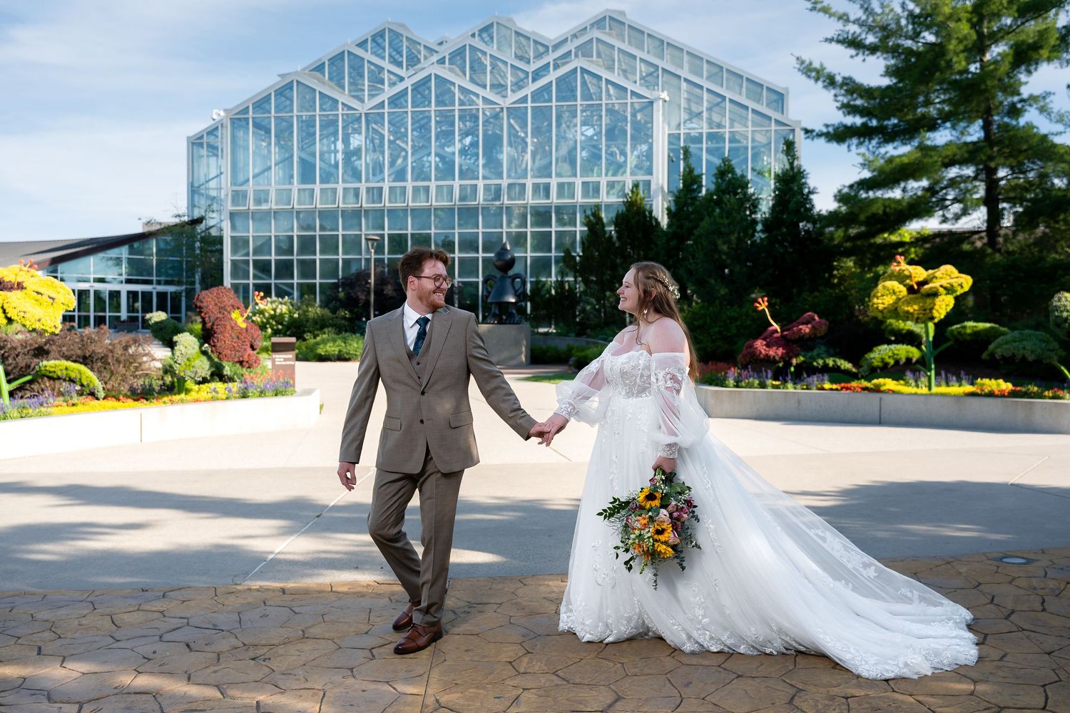Bride and groom holding hands walking in front of a stunning glass greenhouse botanical garden on their wedding day.