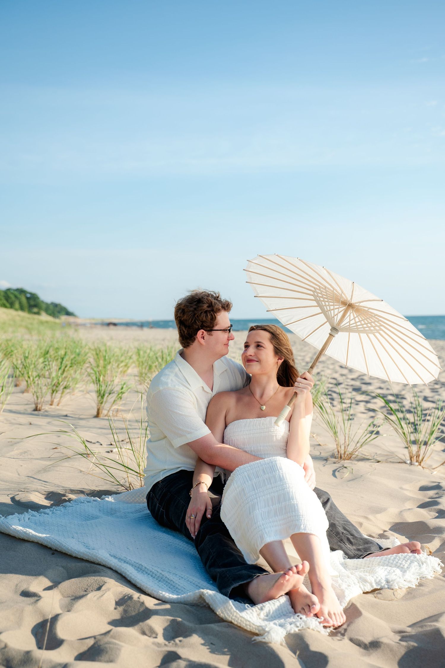 Couple lounging on beach blanket with white parasol against coastal backdrop.