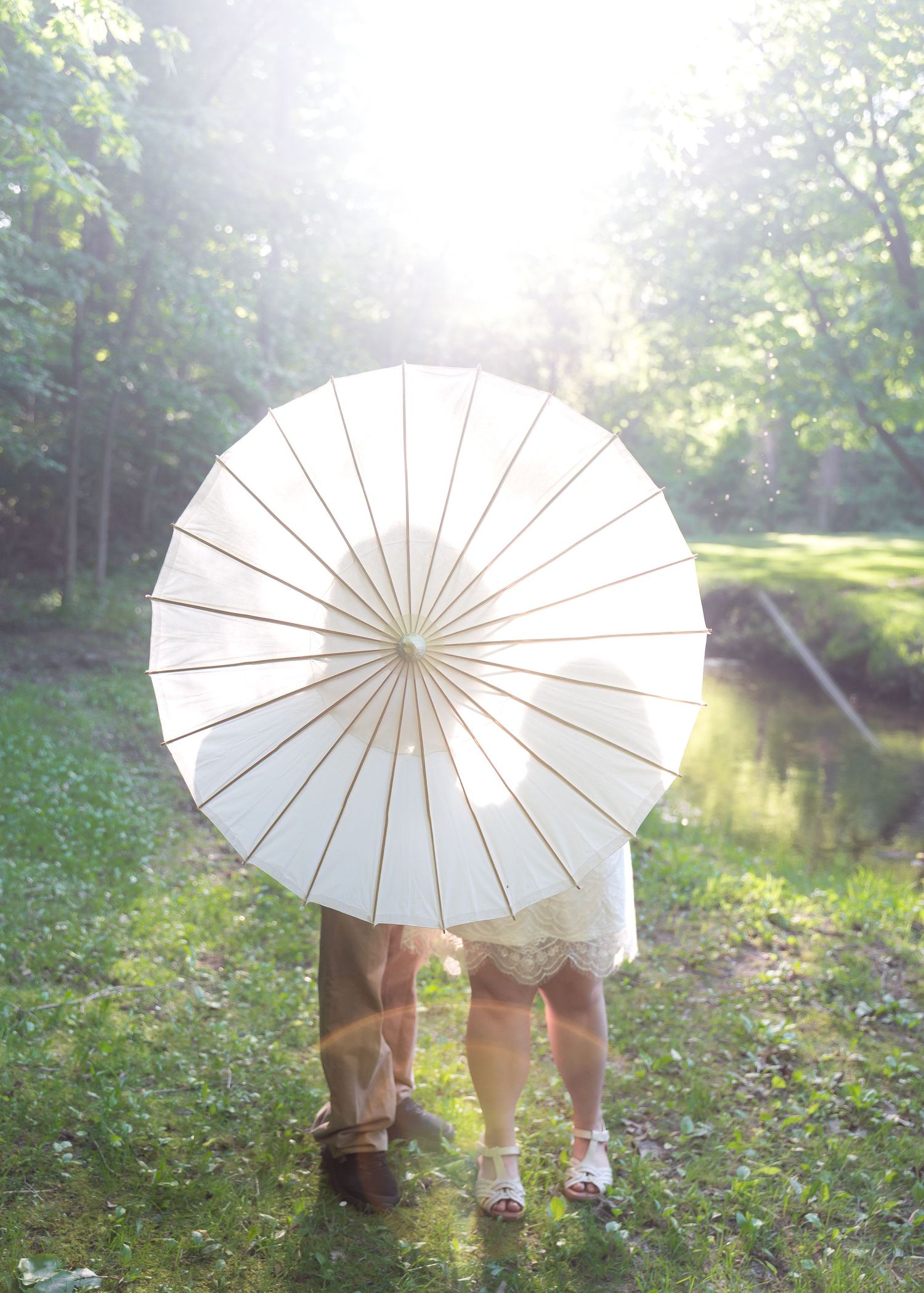 Couple hiding behind a white paper parasol umbrella in a sunlit green garden near a stream.