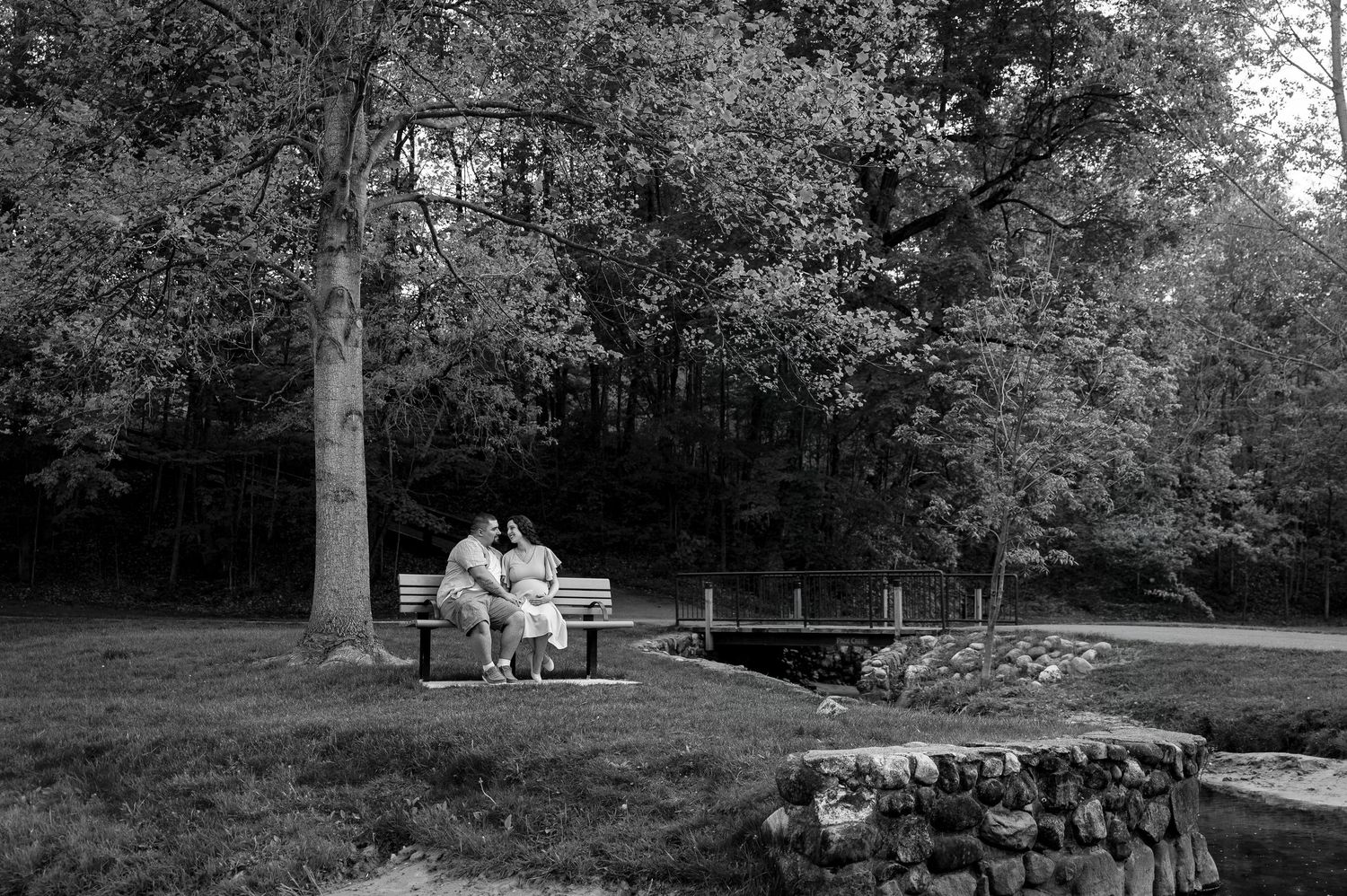 Black and white photo of a couple sitting on a park bench near a stone bridge surrounded by lush trees.