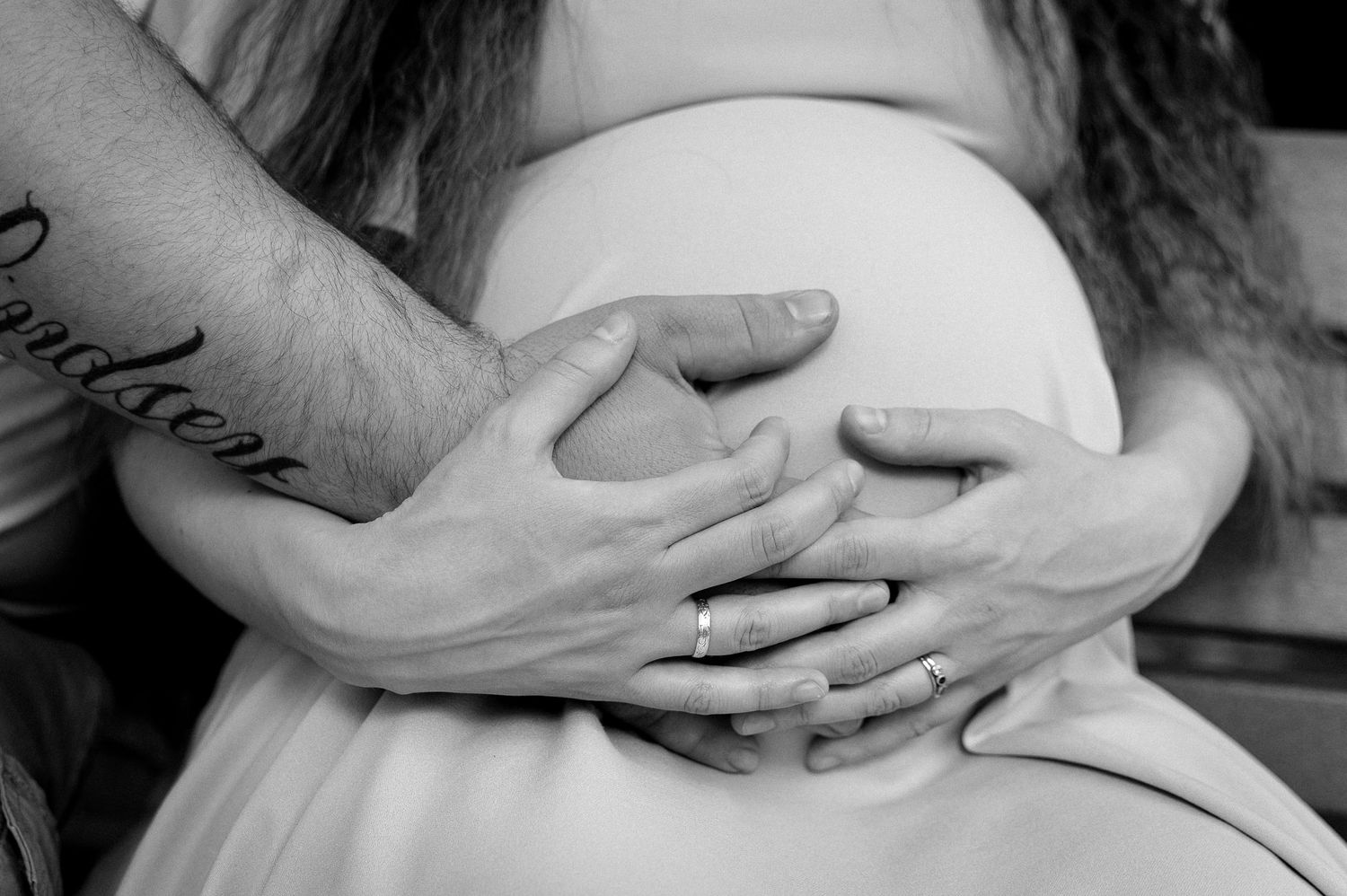 Black and white photo of a pregnant woman with partner's hands gently cradling her baby bump, wearing wedding rings.