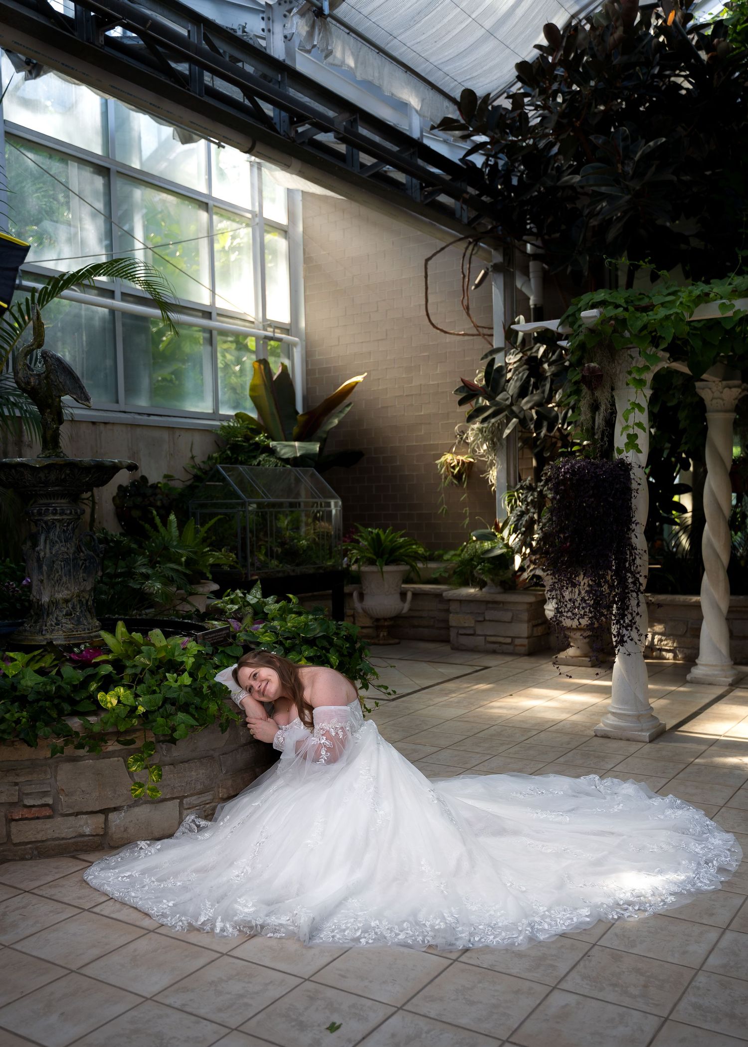 Bride in a flowing white gown poses gracefully on tiled floor inside a lush greenhouse botanical garden.
