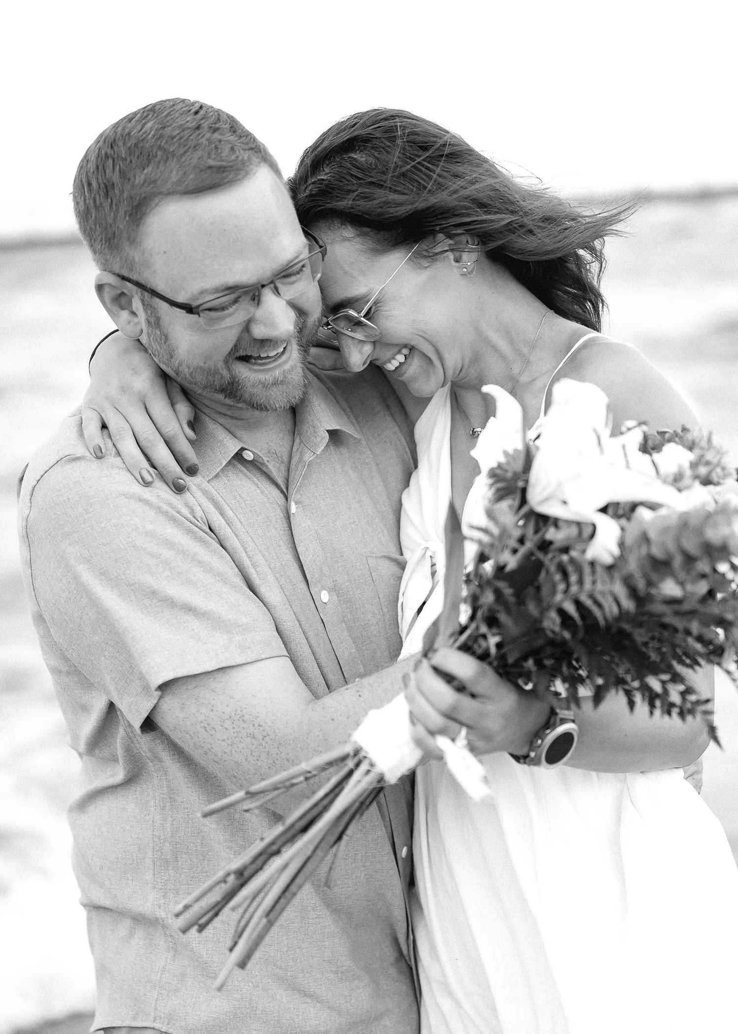 Black and white photo of a laughing couple embracing outdoors, bride holding a bouquet, groom wearing glasses.