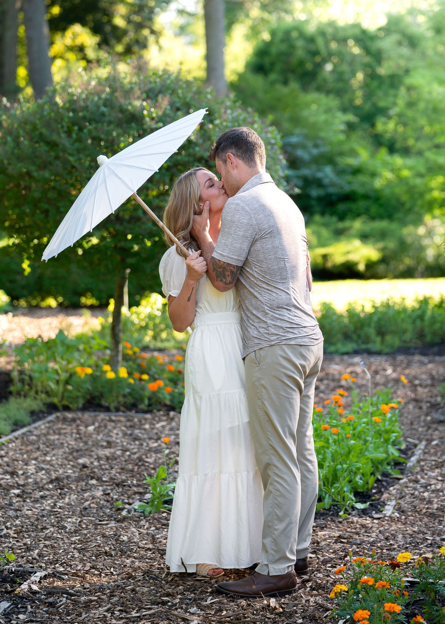 Couple kissing in a garden, woman in white dress holding a white parasol, surrounded by colorful flowers and greenery.