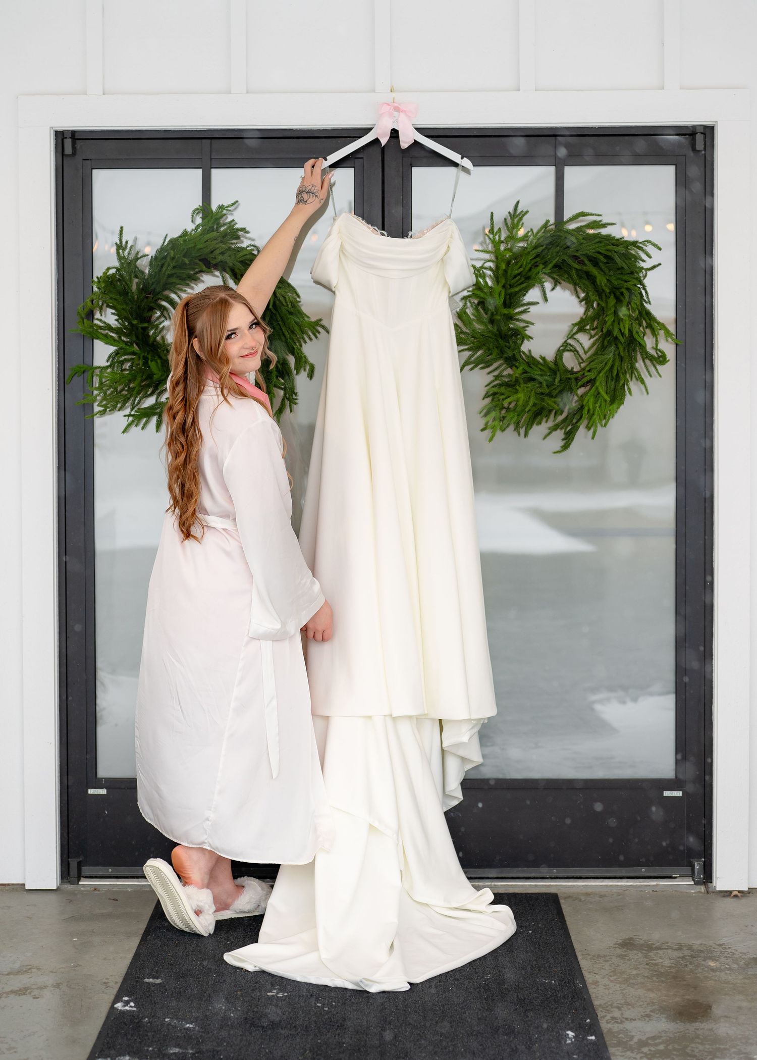 Bride in white robe holding up elegant wedding dress on hanger, framed by green wreaths on glass doors.