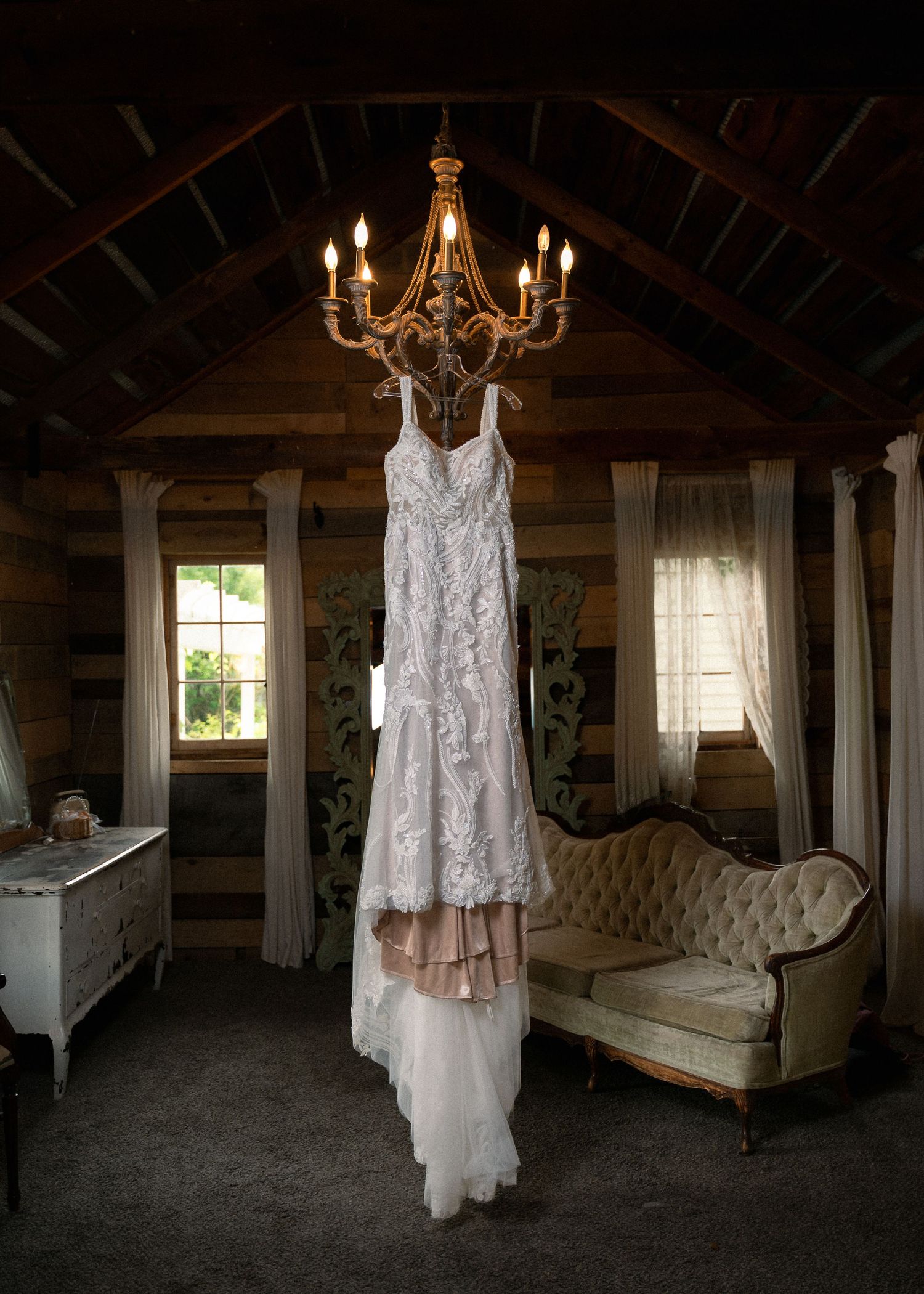 A lace wedding dress hanging from a rustic chandelier in a cozy log cabin bridal suite with vintage furniture.