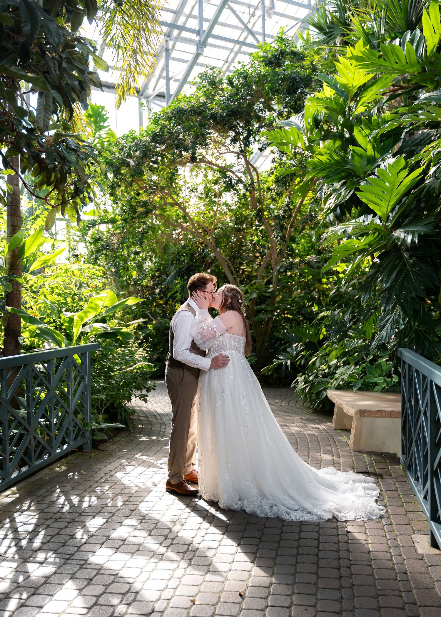 Bride and groom share a kiss in a lush tropical greenhouse garden, bride in elegant white gown with long train.
