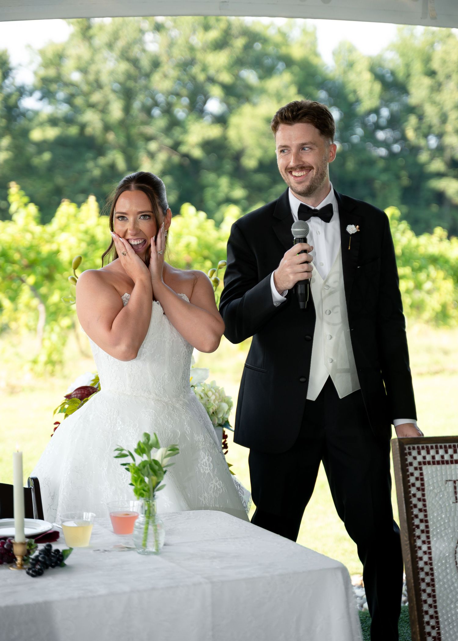 Bride and groom smiling during wedding reception speech at outdoor vineyard venue, bride in white gown, groom in black tuxedo.
