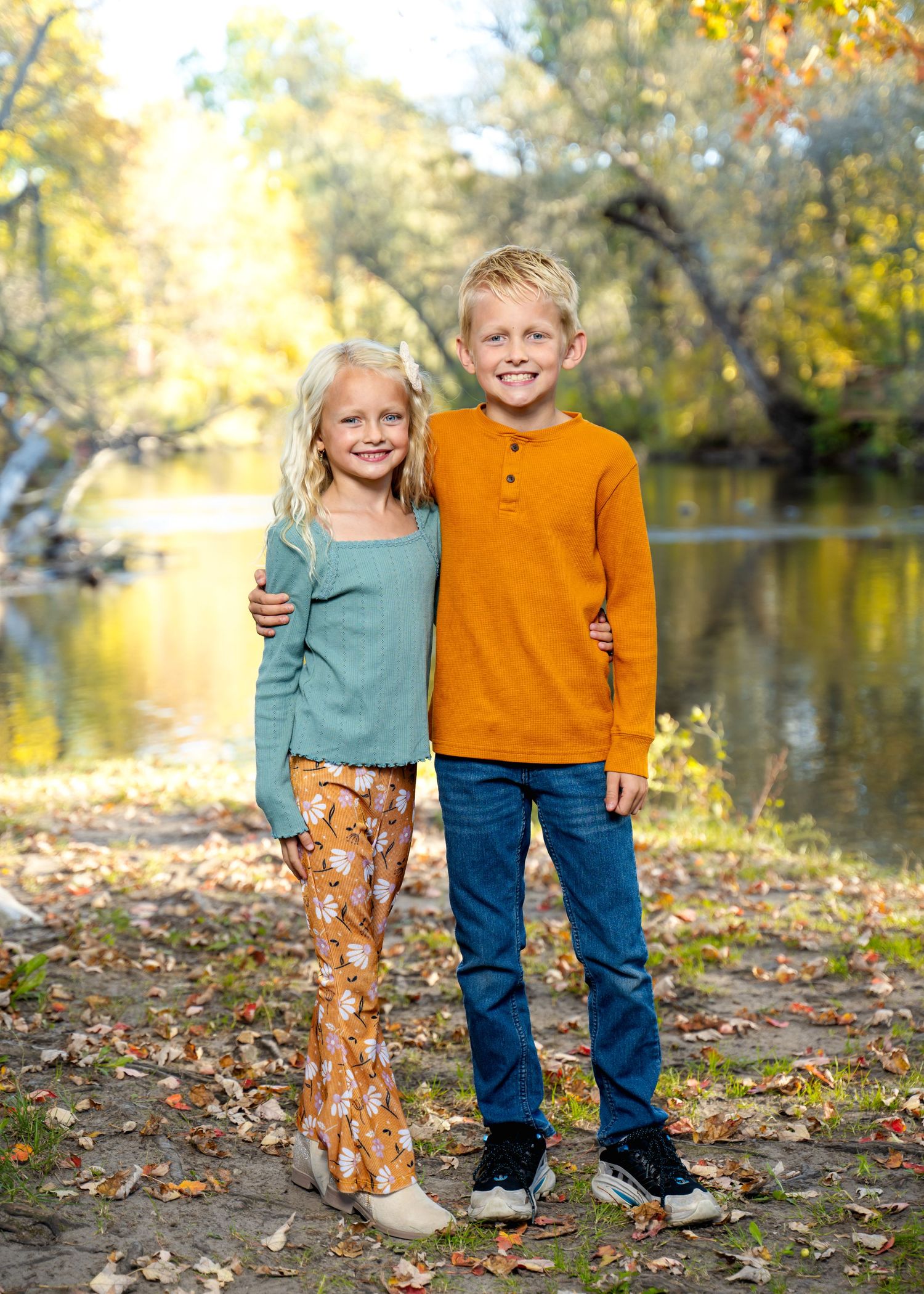 Two children stand together by a lake on an autumn day with golden trees in the background.