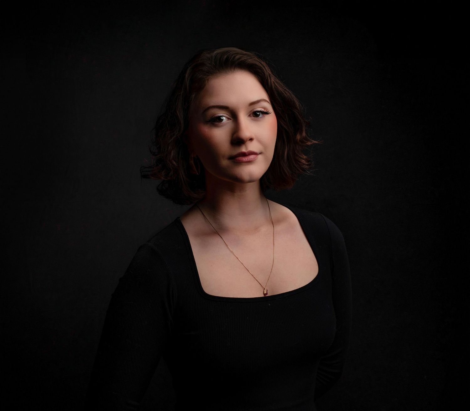 A dramatic portrait against a dark background featuring a black top and simple necklace.