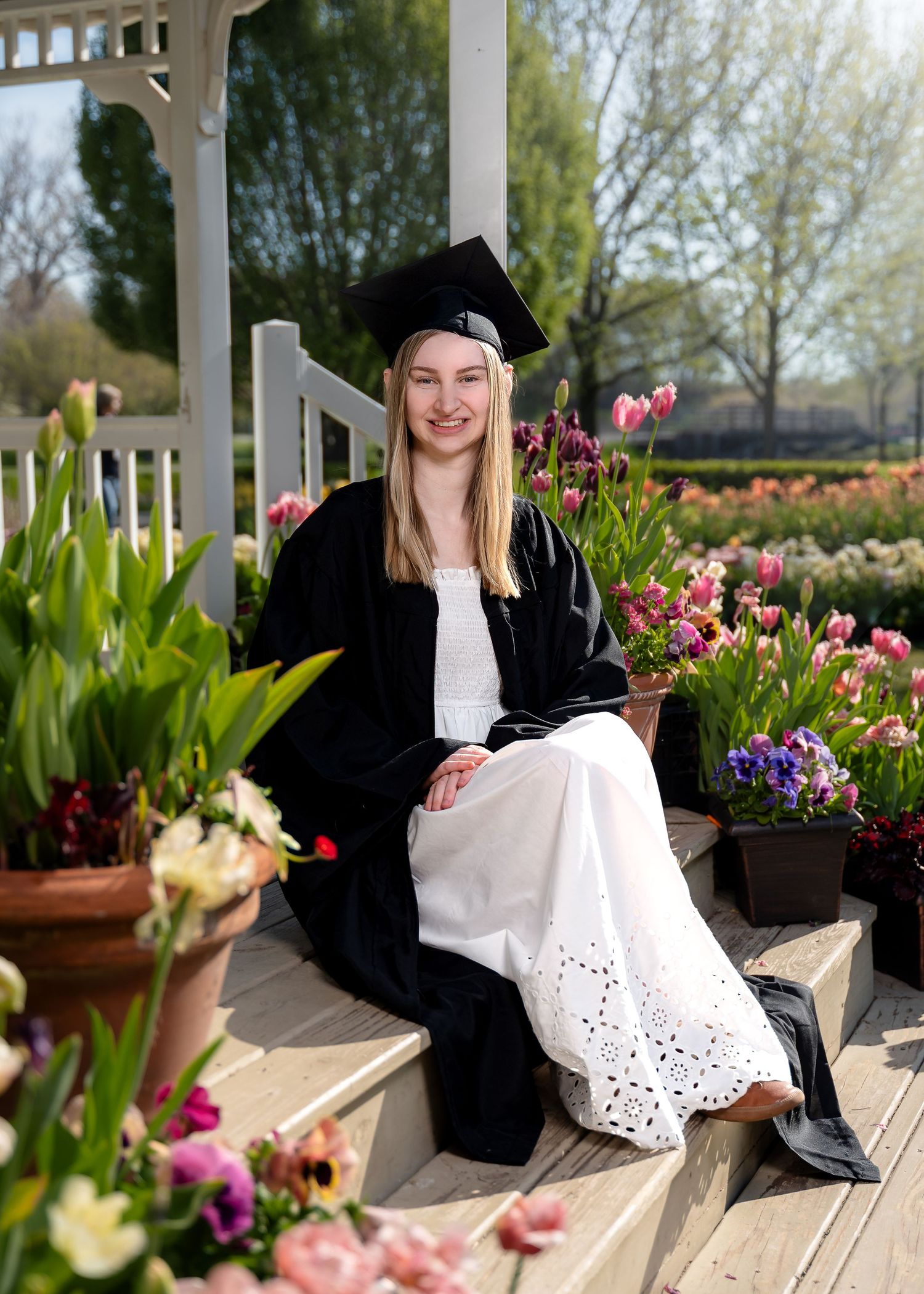 Young woman in black graduation cap and gown sits among colorful spring tulips on gazebo steps, smiling.
