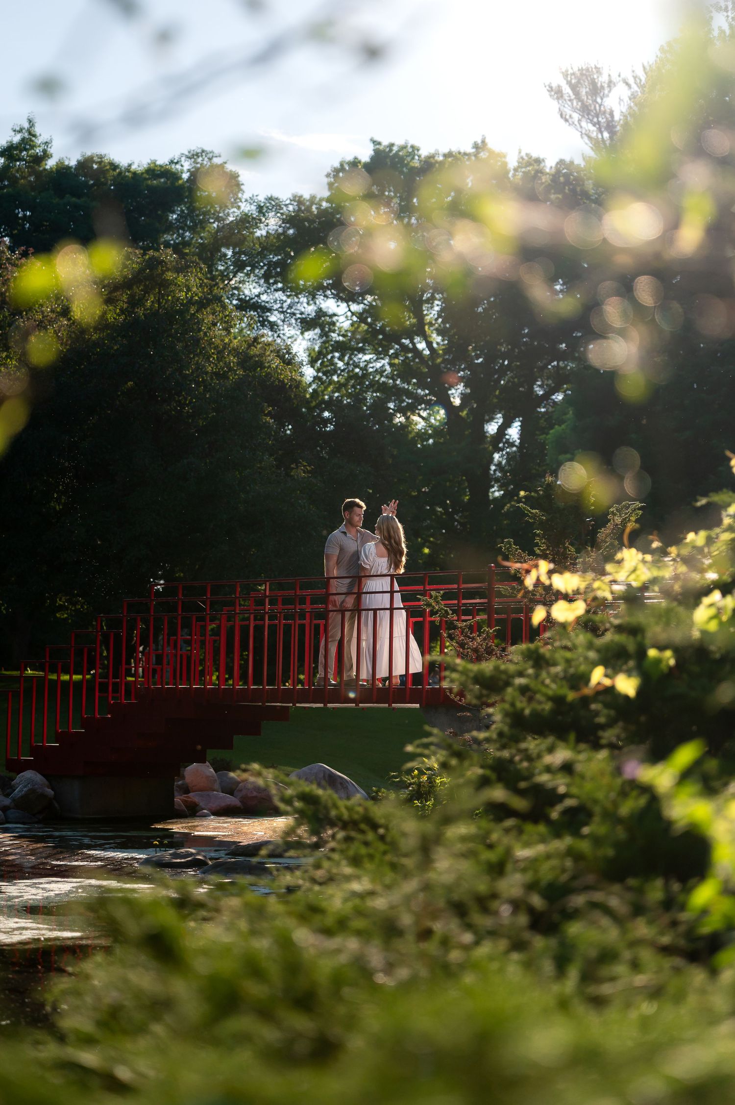 Couple in wedding attire standing on a red bridge surrounded by lush green trees in a romantic garden setting.