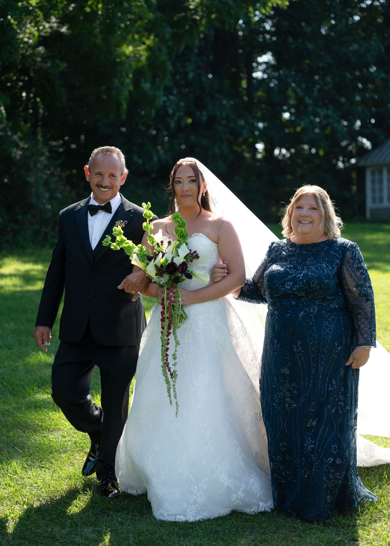 Bride in white gown holding bouquet walks with parents in formal attire on green lawn at outdoor wedding.