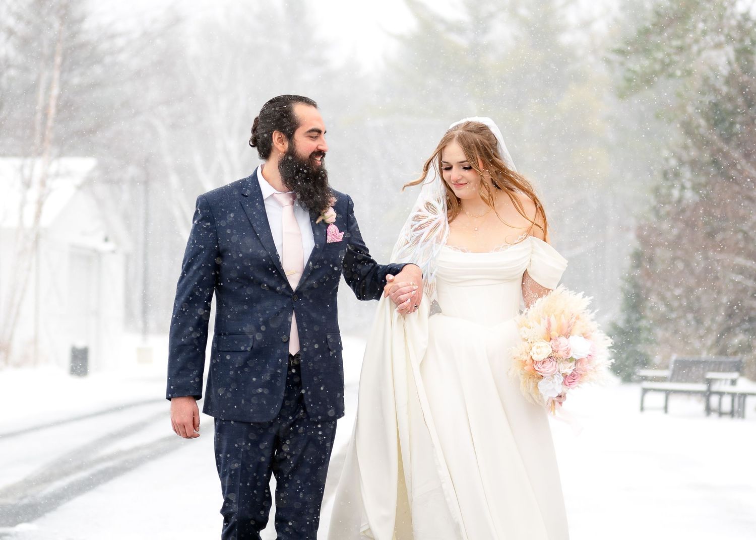 Bride and groom walking hand in hand in a snowy winter wonderland, bride holding a pastel bouquet in elegant white gown.