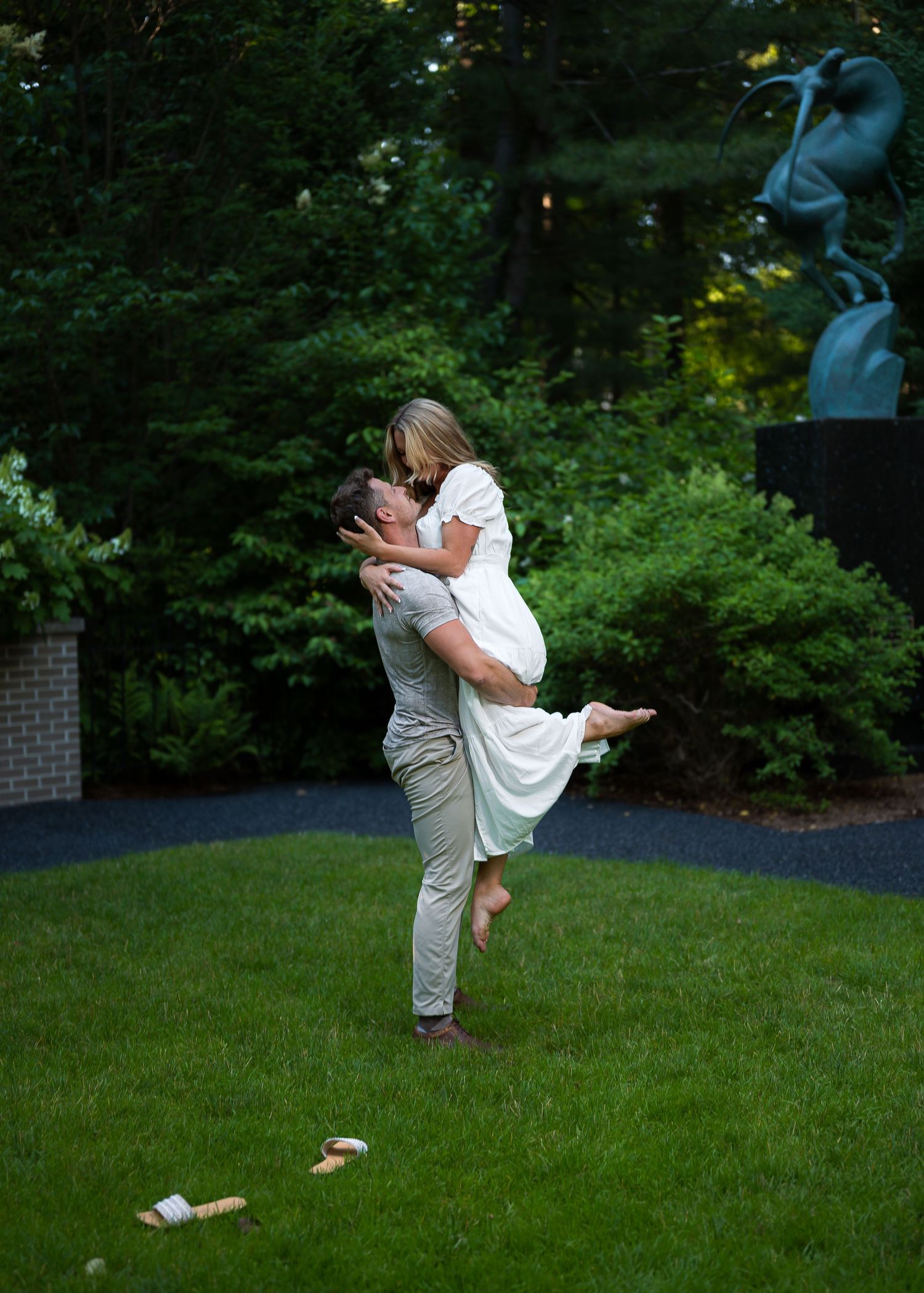 Man lifting woman in white dress for a kiss outdoors on green lawn with lush trees and bronze sculpture backdrop.