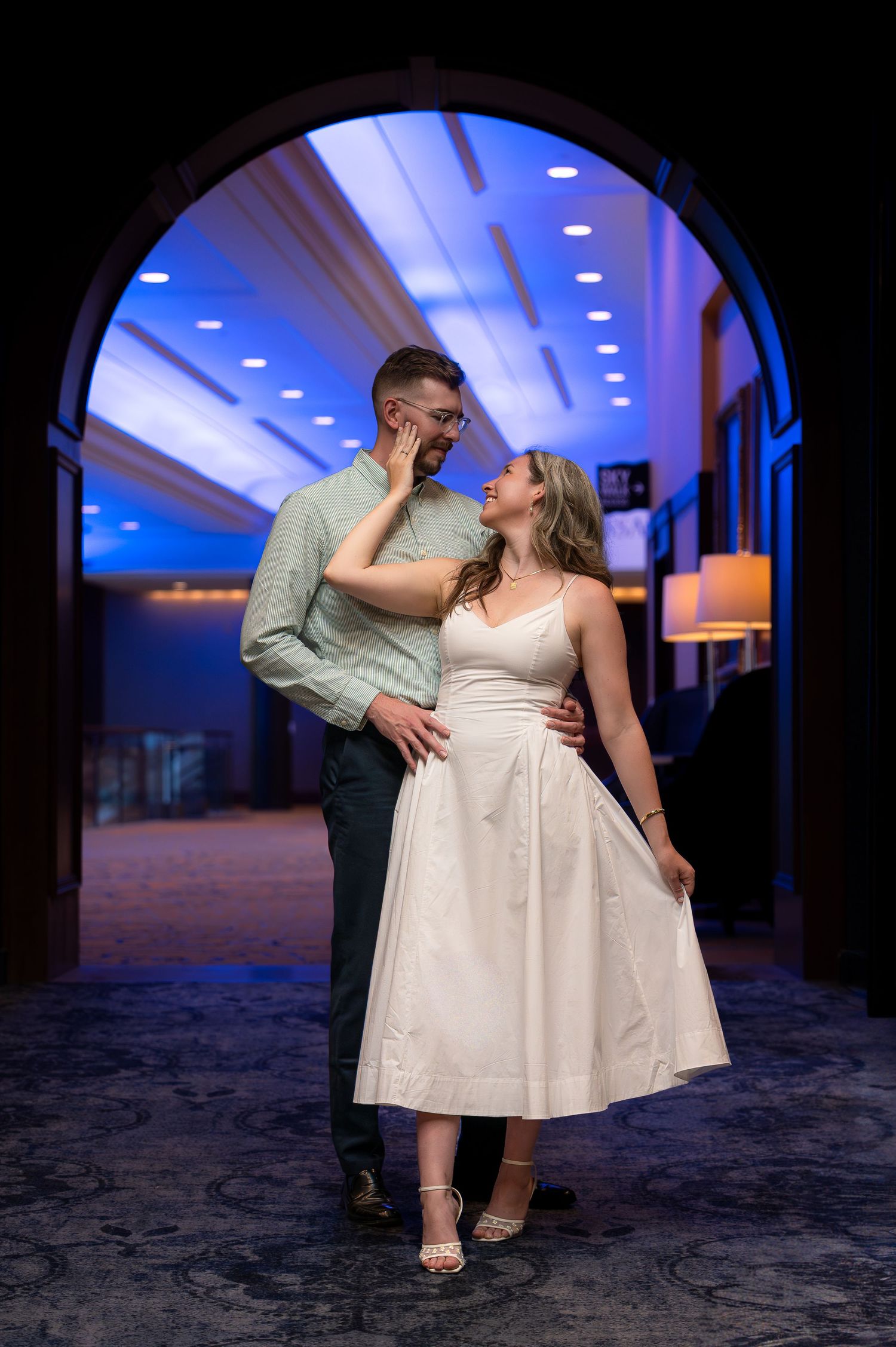Couple embracing under an elegant arch in a hotel corridor, woman in white dress gazing up at man in blue ambient lighting.