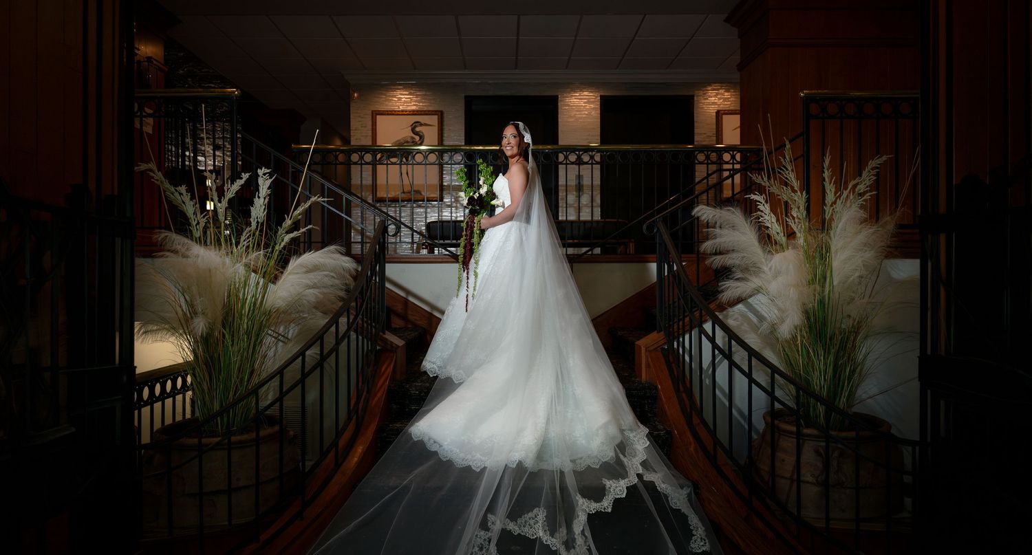 Bride in elegant white gown and long veil stands on grand staircase in a beautifully lit wedding venue.