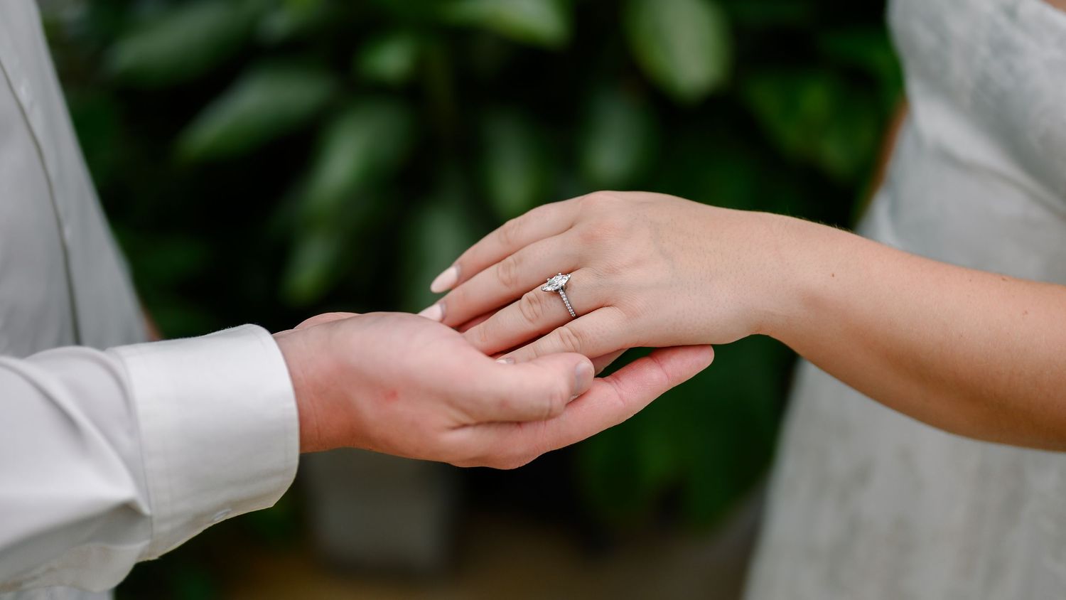 Close up of two hands touching during wedding ring exchange with green foliage in background.
