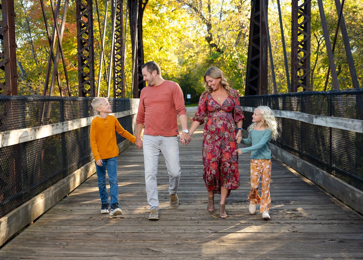 Family walking together on wooden bridge surrounded by autumn foliage and golden sunlight.