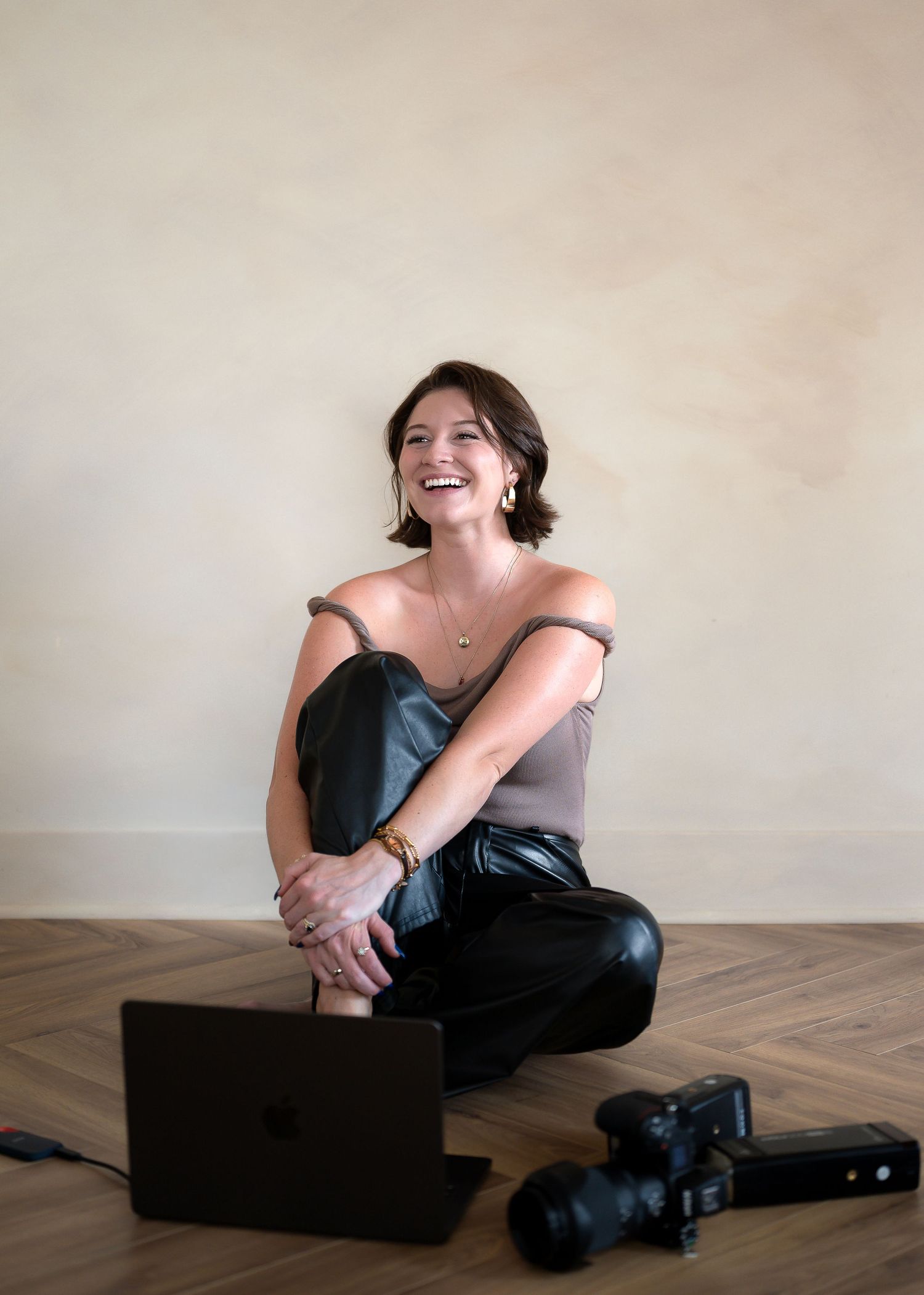 Smiling woman sitting on floor with laptop and camera, wearing gray top and black leather pants.