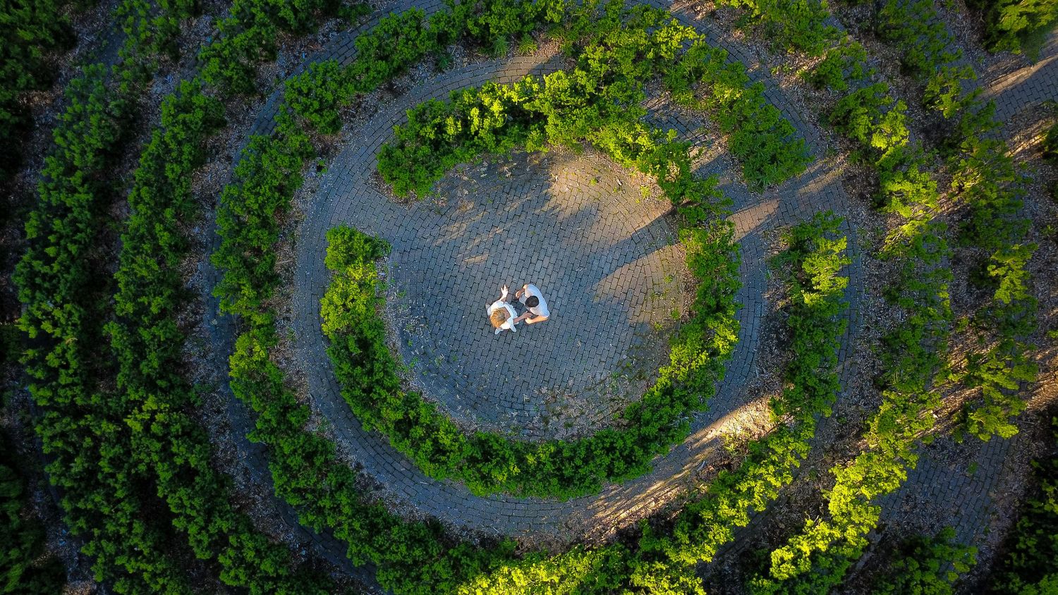 Aerial view of two people sitting in a circular gravel clearing surrounded by lush green spiral hedges.