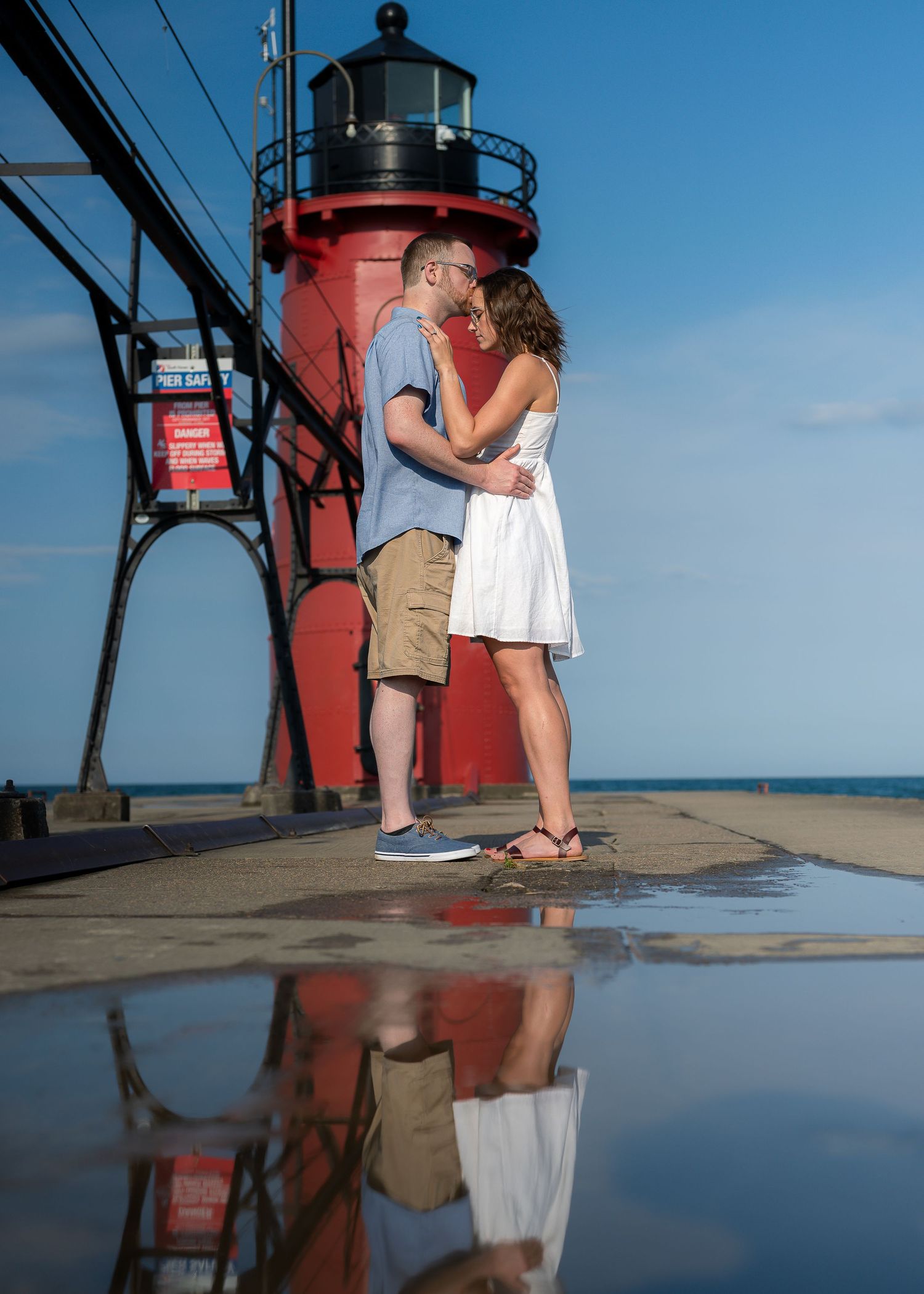 Couple embracing and kissing near a red lighthouse, with their reflection visible in a puddle below on a sunny day.