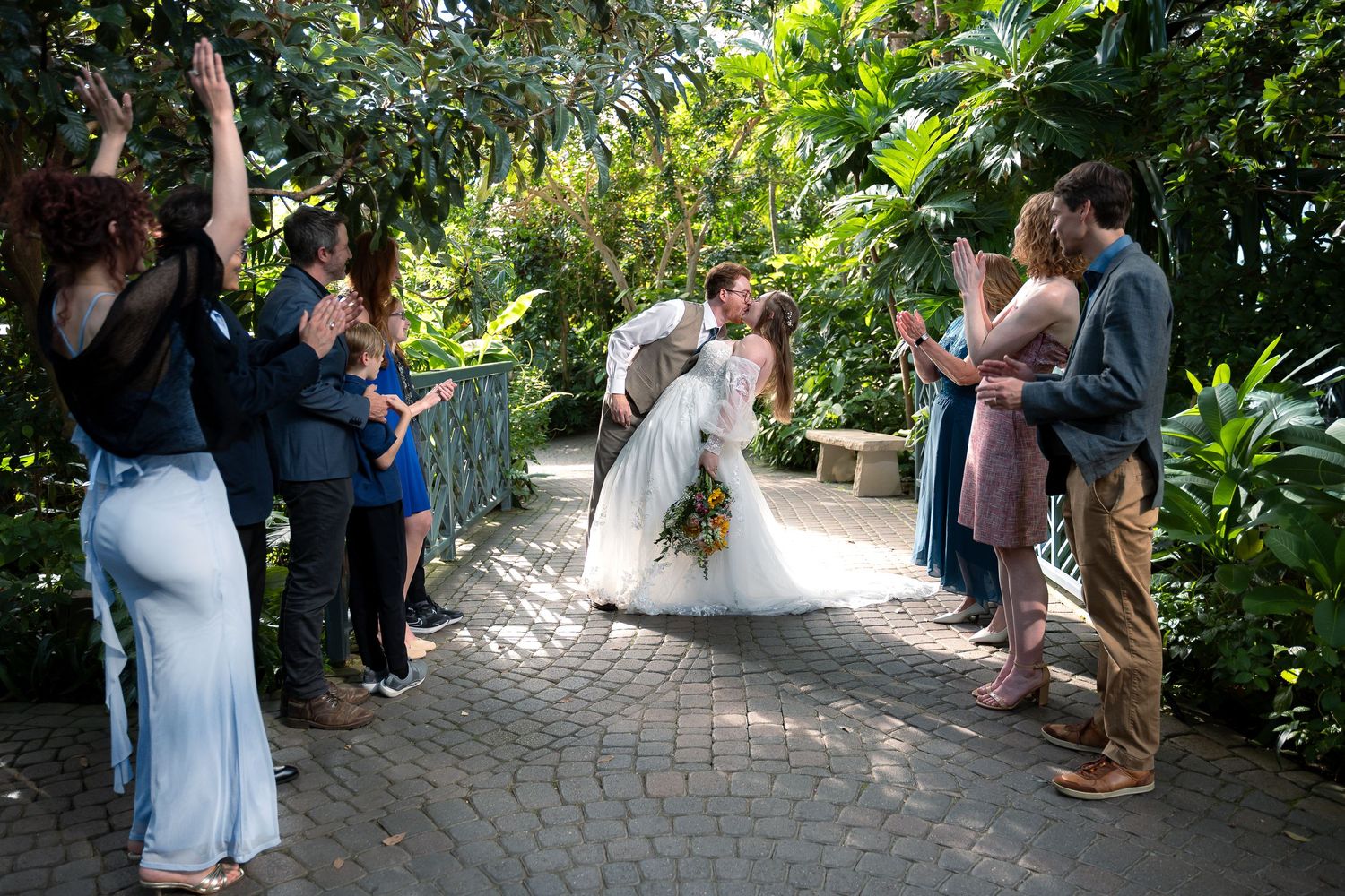 Bride and groom share a kiss on a garden path surrounded by cheering guests at an outdoor wedding ceremony.