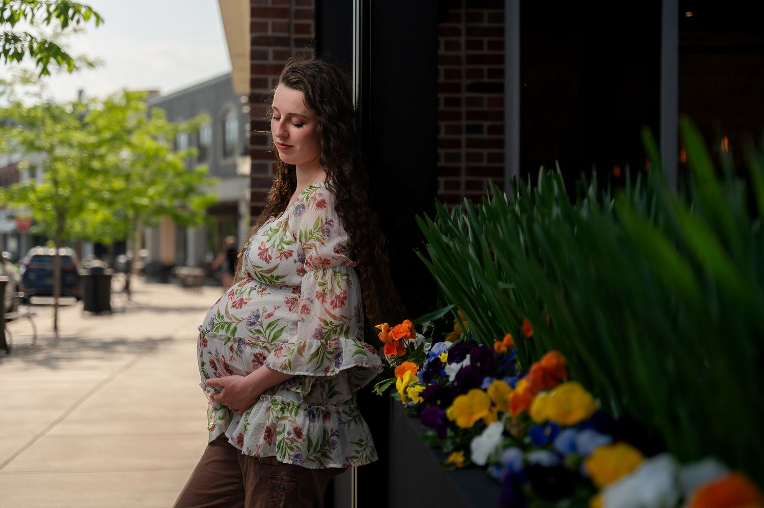 Pregnant woman in floral top cradling baby bump beside colorful flowers on a sunny urban sidewalk.