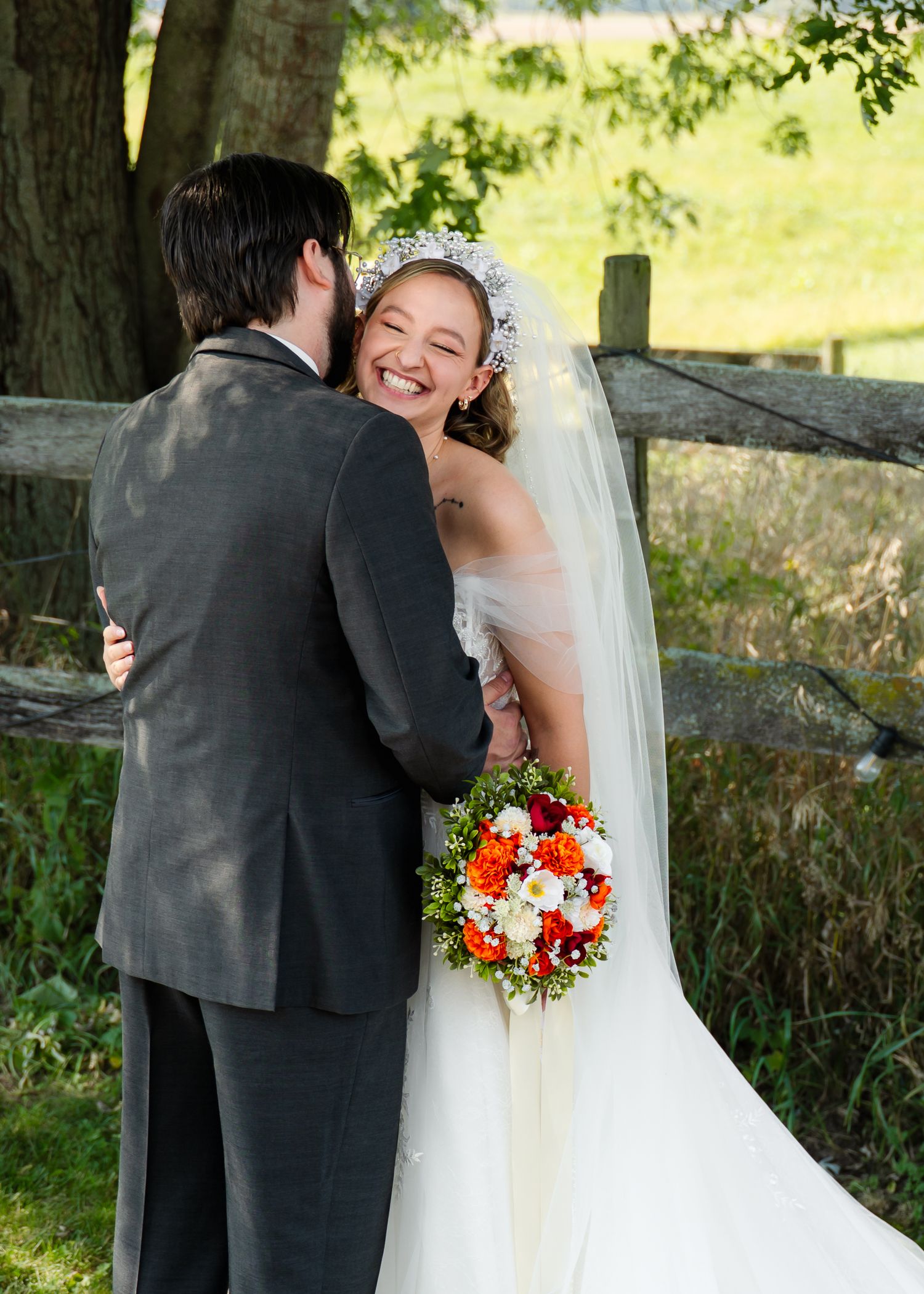 Joyful wedding couple embraces by a rustic wooden fence in natural outdoor setting.