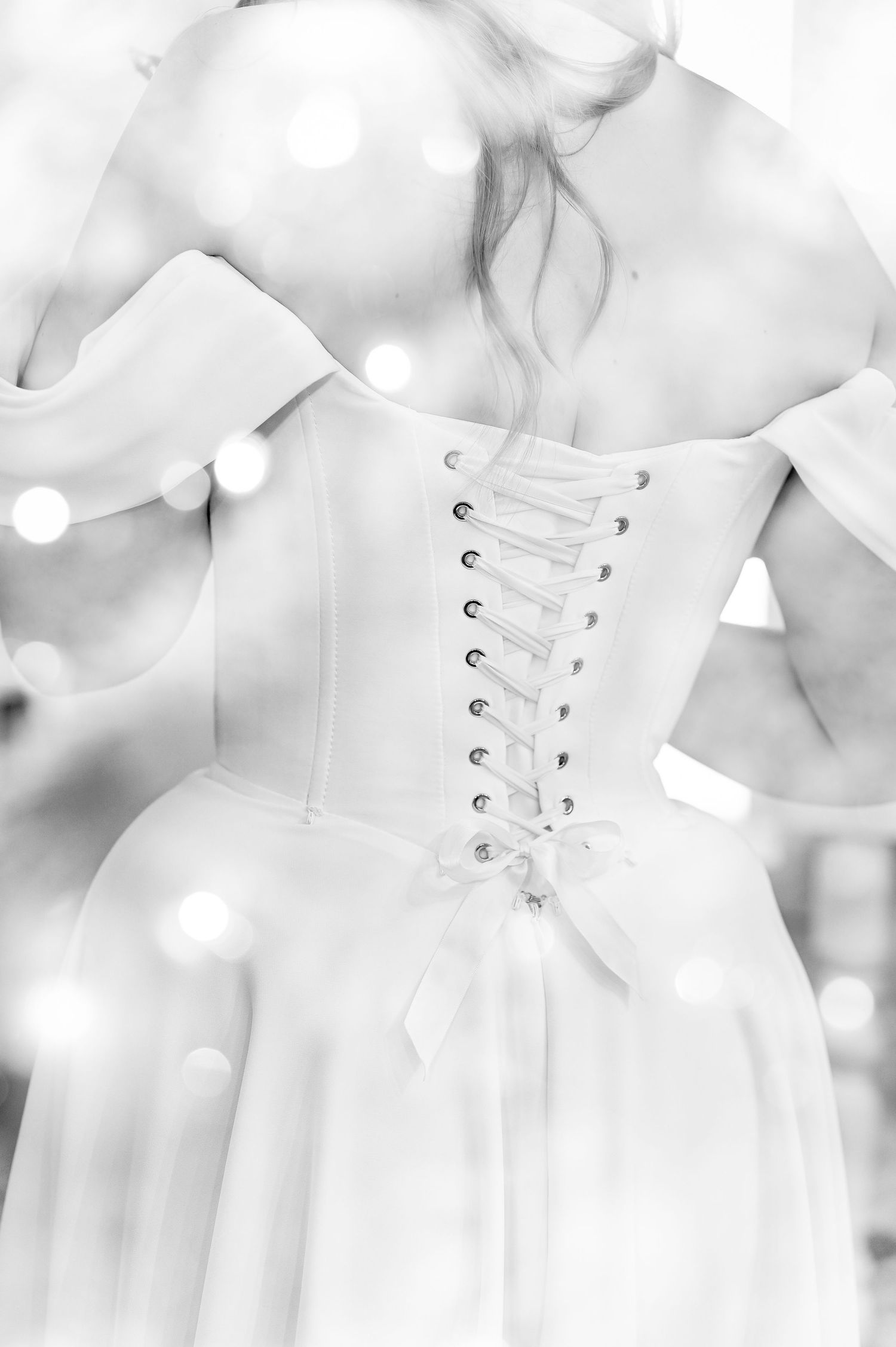 Black and white photo of a bride in a corset-back wedding gown, showcasing elegant lace-up detailing with bokeh lights.