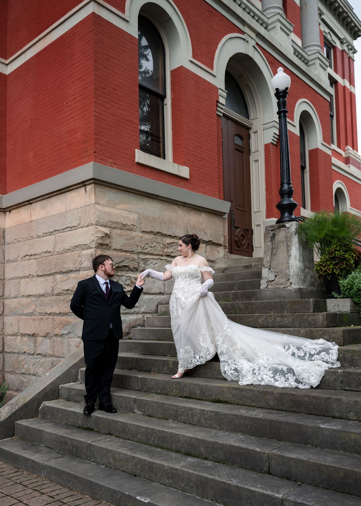 Bride and groom hold hands on stone steps outside a historic red brick building during their wedding day.