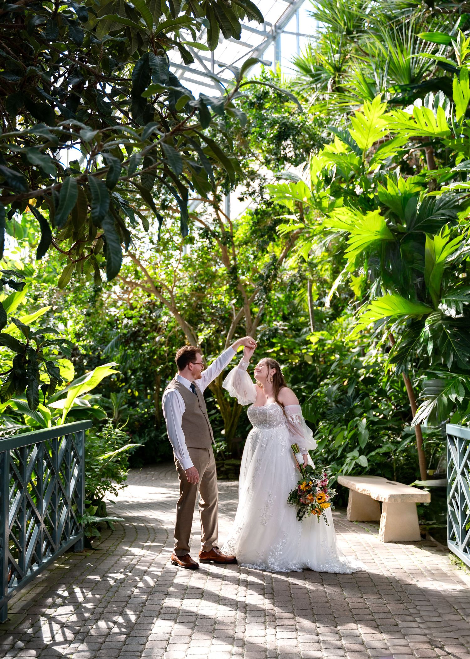 Bride and groom dancing in a lush tropical greenhouse conservatory, bride holding colorful bouquet in white gown.