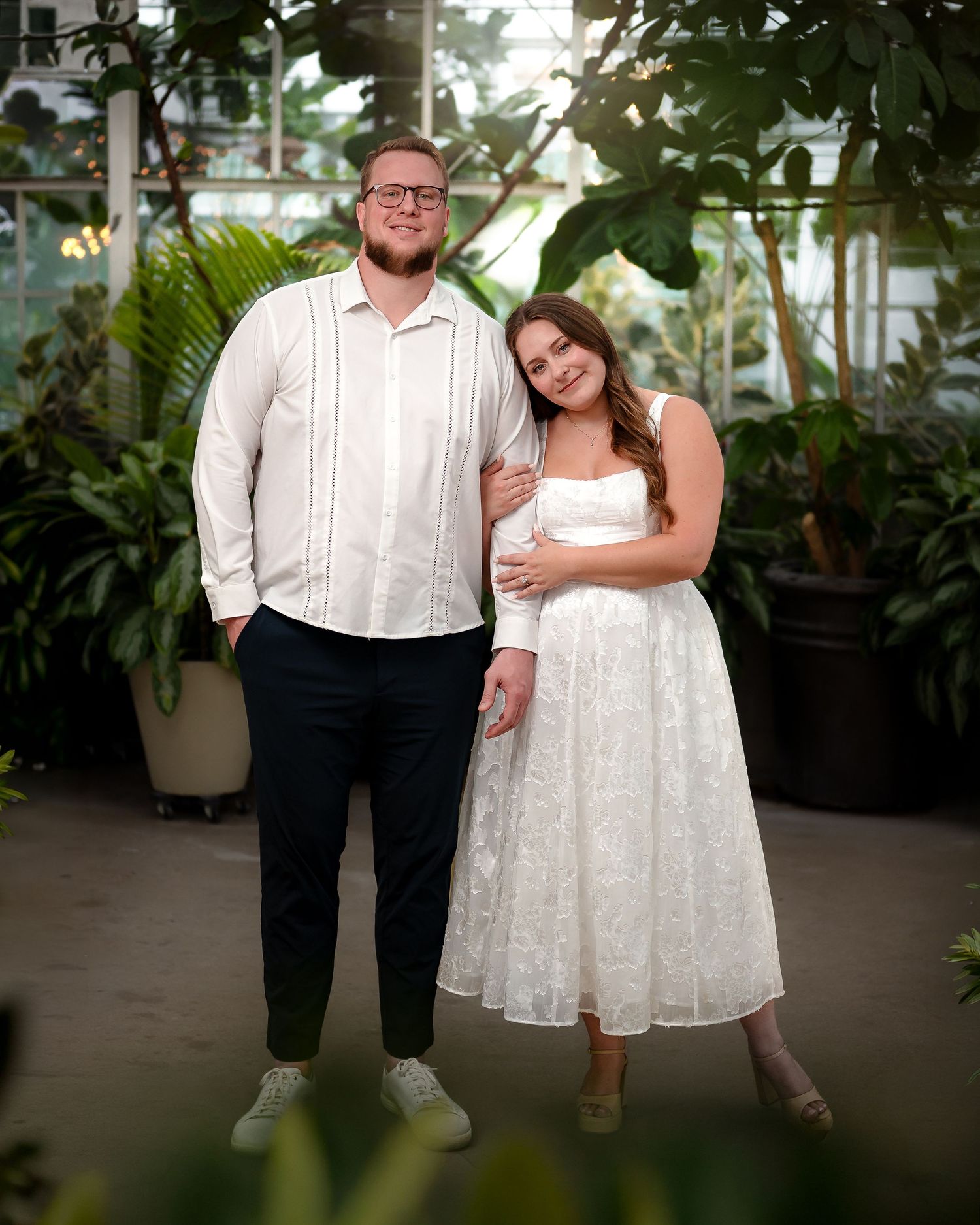 A couple poses together in a greenhouse setting surrounded by lush greenery and tropical plants.