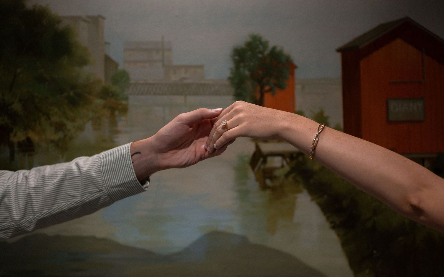 Two hands gently touching fingertips near a serene river with a red building in the background.