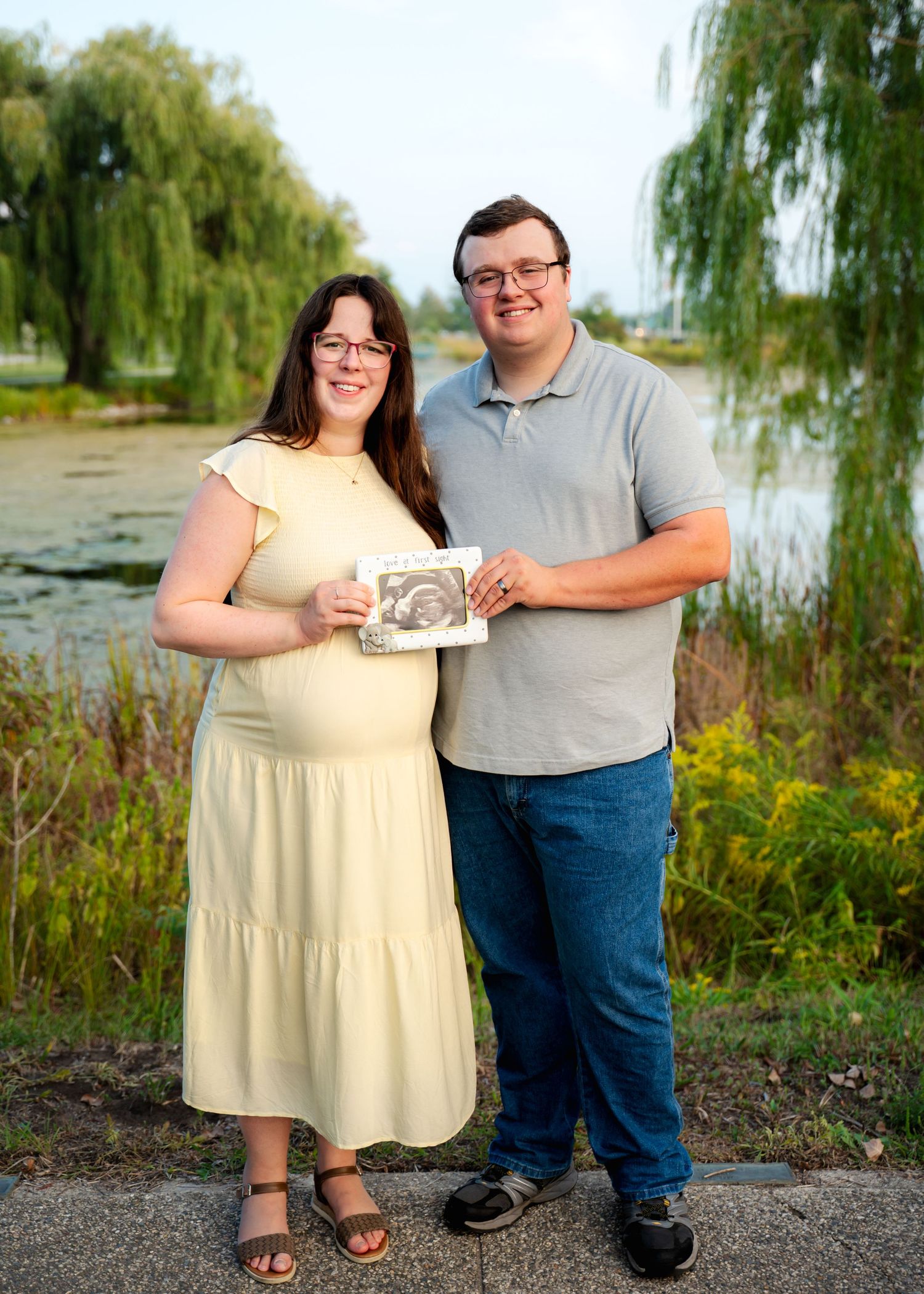 Couple holding ultrasound photo by willow tree near waterfront.