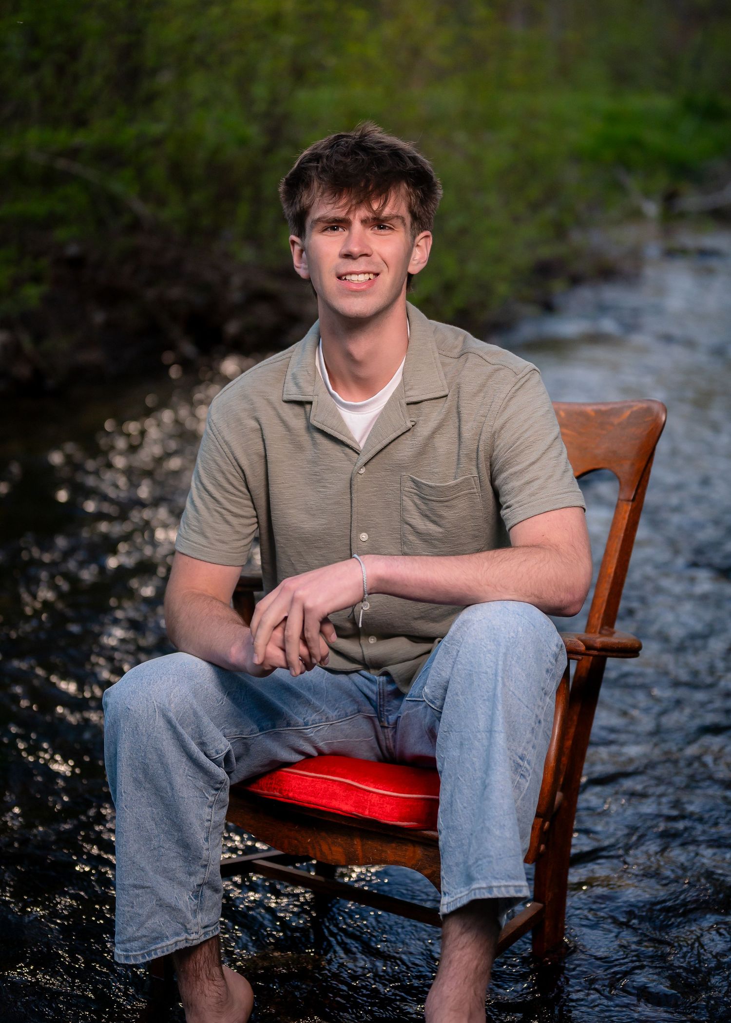 Young man sitting barefoot on a red-cushioned wooden chair in a shallow creek, wearing a gray shirt and jeans.
