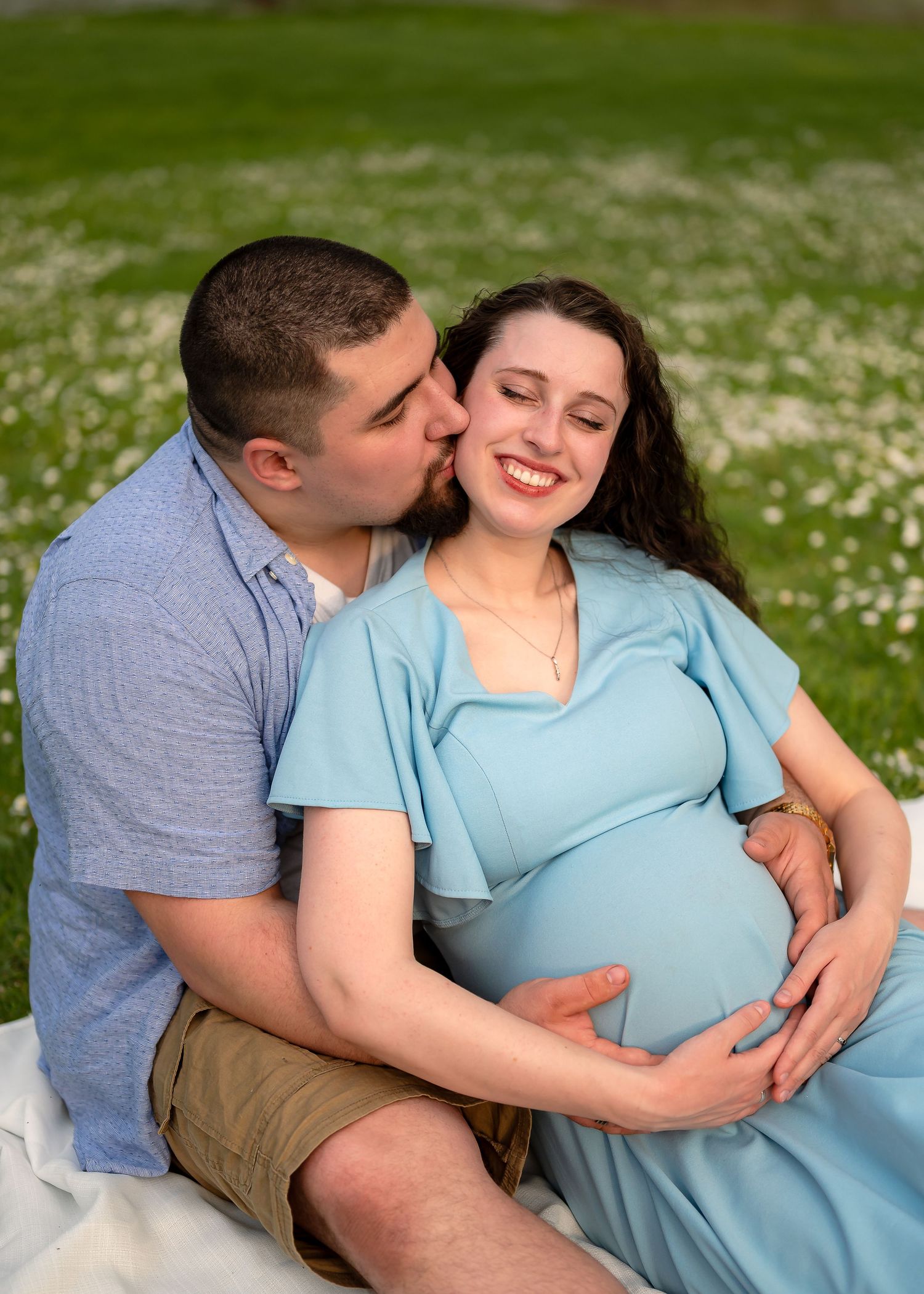 A couple sits in a flower field, man kissing pregnant woman's cheek as she smiles, both cradling her baby bump.